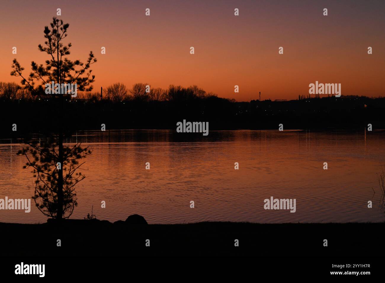 Sullo sfondo di un lago profondo, un tramonto arancione si alza nel cielo, le onde luminose sulla superficie dell'acqua riflettono colori caldi e sagome Foto Stock