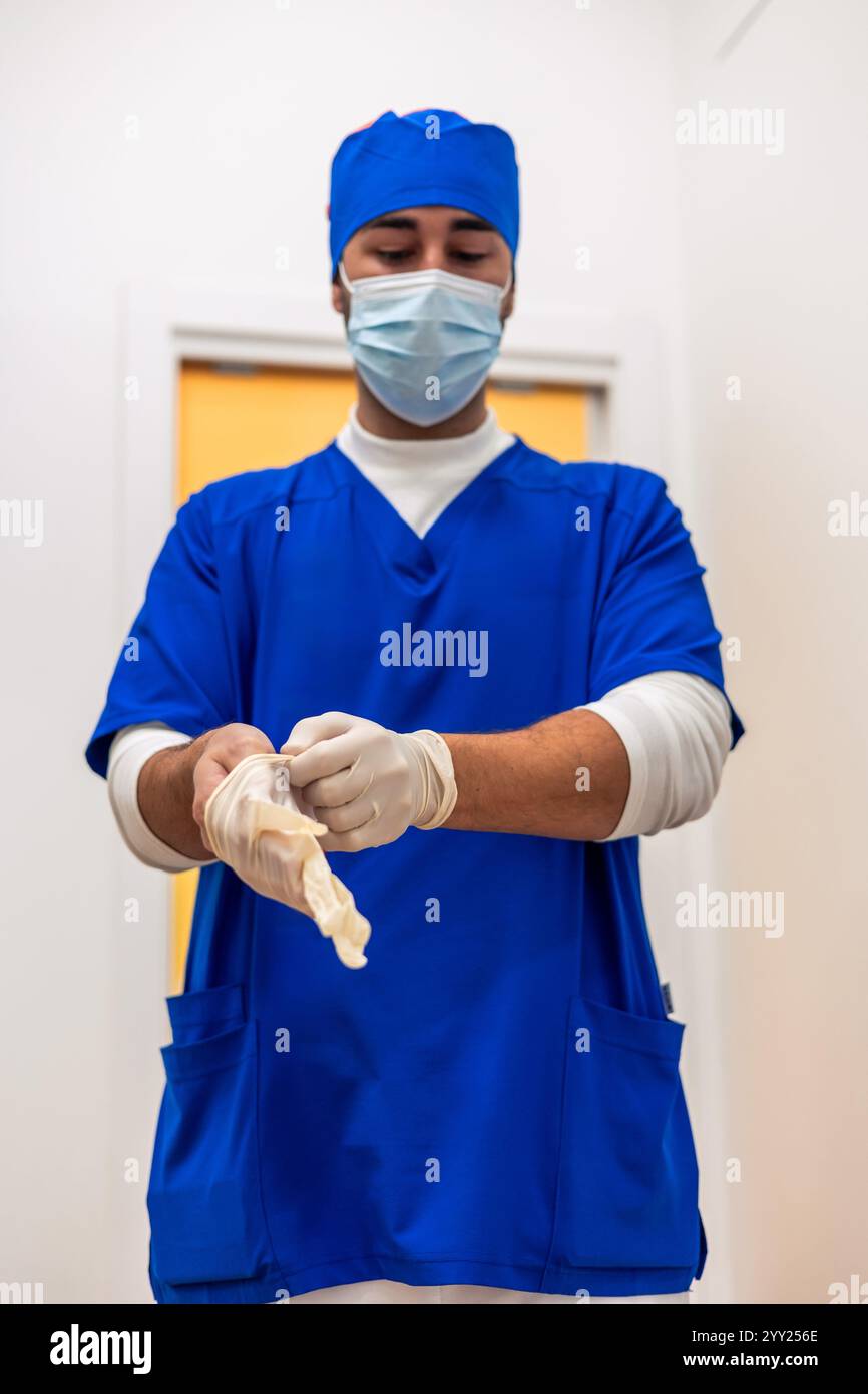 Giovane dentista maschio che indossa uniforme chirurgica blu, cappuccio e maschera indossando guanti sterili in lattice, preparandosi per l'intervento chirurgico all'interno della clinica odontoiatrica Foto Stock