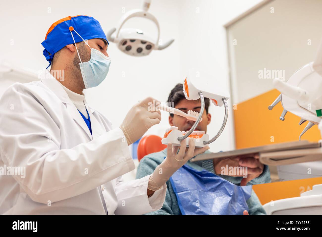 Dentista che indossa maschera e guanti che mostrano come spazzolare correttamente i denti al suo paziente utilizzando un modello di dentatura e uno spazzolino da denti in una clinica odontoiatrica Foto Stock