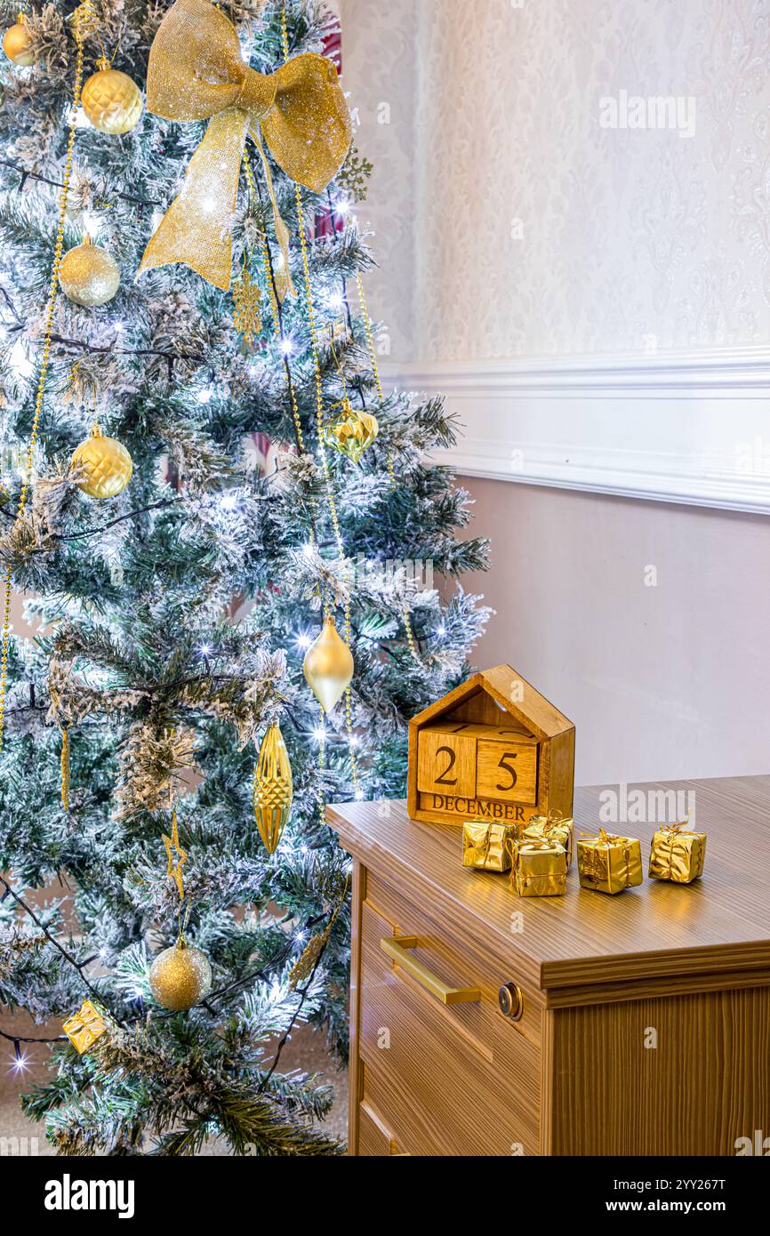 Albero di Natale con decorazioni in oro e luci scintillanti accanto a un calendario in legno datato 25 dicembre, sopra un armadietto di archiviazione in legno per ufficio Foto Stock