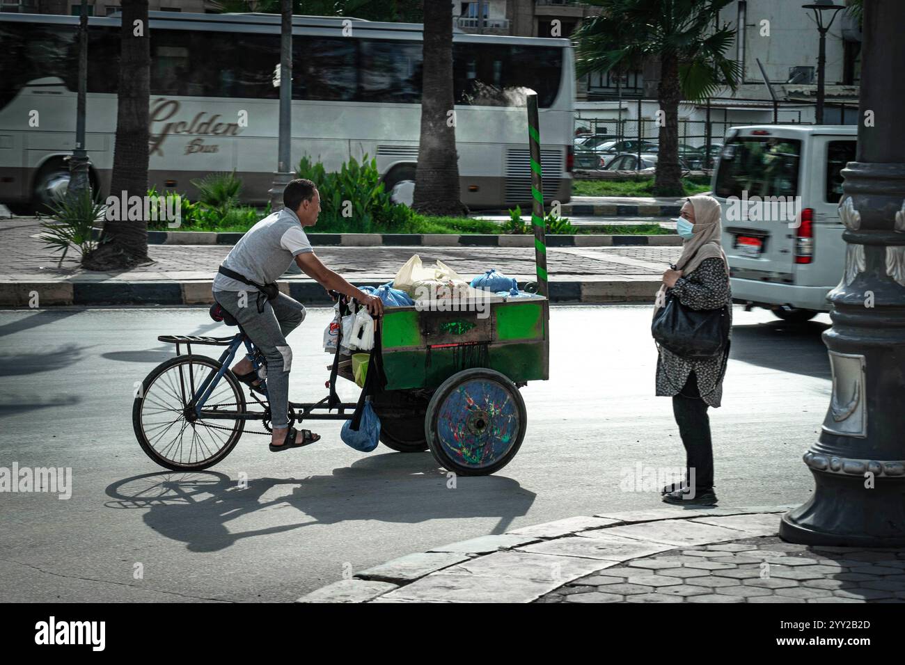 Una scena di strada ad Alessandria, in Egitto. Un uomo guida una bicicletta con un carrello mobile dotato di fornello, borse e provviste, mentre una donna è nelle vicinanze. Foto Stock