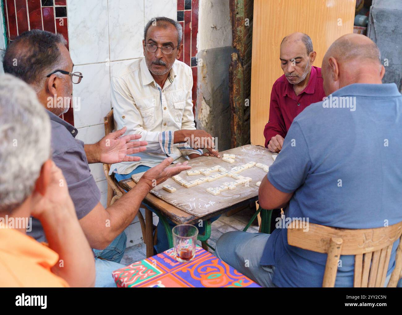 Gruppo di anziani che giocano a domino in un rustico cafй all'aperto ad Alessandria, Egitto. Una scena di strada calda e sociale che cattura la vita quotidiana locale. Foto Stock