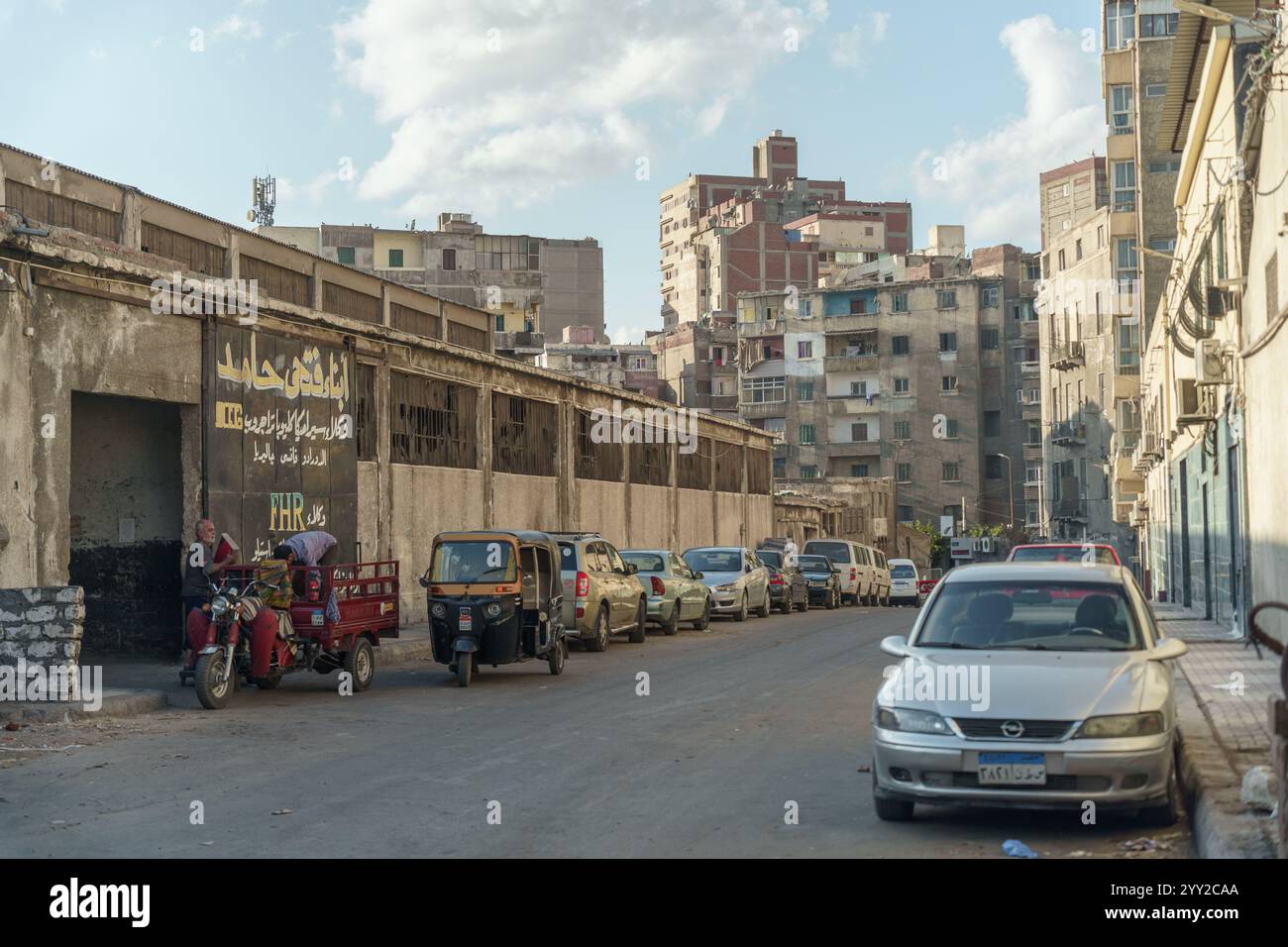 Scena urbana di Alessandria, Egitto, con auto parcheggiate, tuk-tuk e vecchi edifici industriali sotto un cielo limpido. Una miscela di vita quotidiana e decadimento. Foto Stock