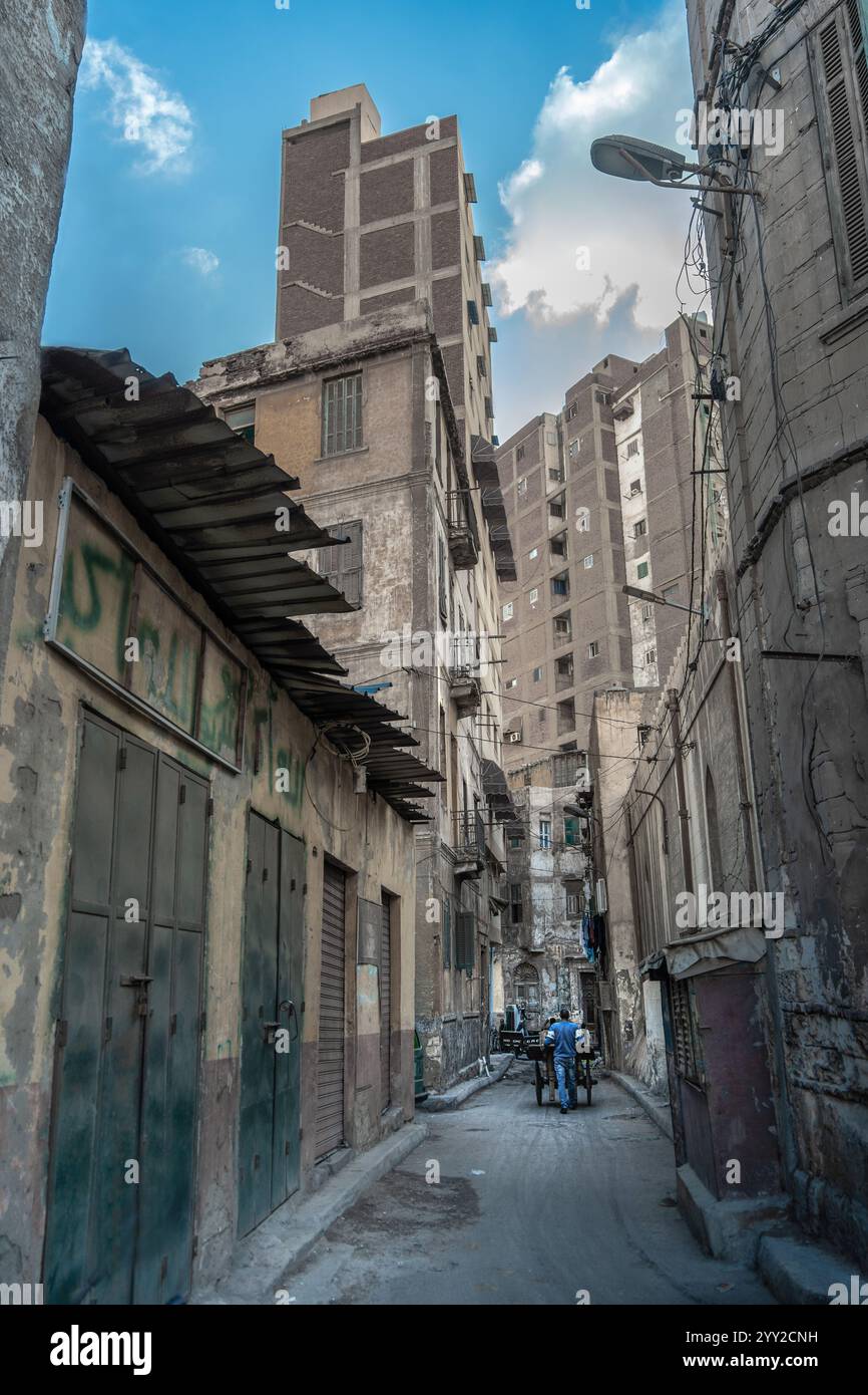 Vicolo stretto di Alessandria, Egitto, circondato da edifici intemprati, muri di graffiti e vivace vita urbana con la gente del posto che spinge i carri sotto il cielo Foto Stock