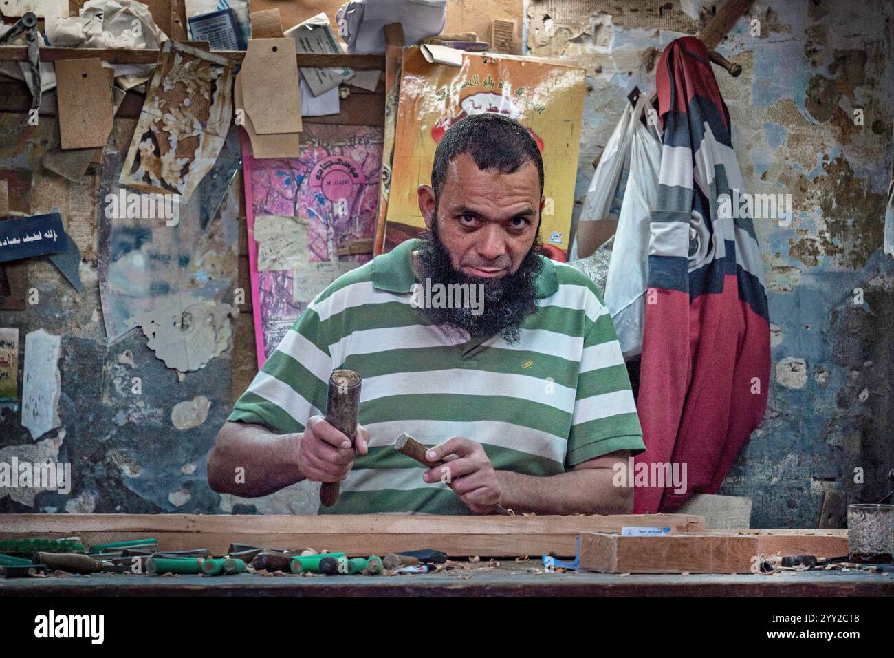 Un artigiano con una camicia verde a righe lavora intensamente con gli attrezzi in un laboratorio pieno di legno fatto a mano e pareti rustiche ad Alessandria, in Egitto. Foto Stock