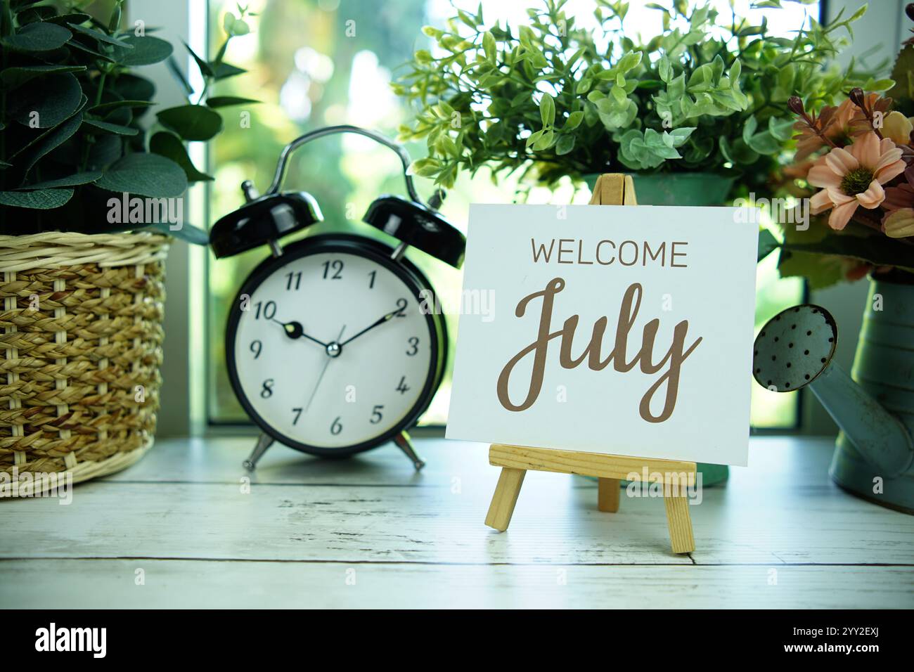 Testo di benvenuto di luglio su carta con cavalletto e sveglia sullo sfondo della finestra Foto Stock