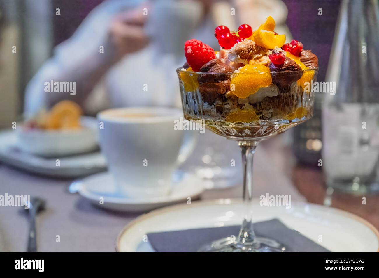 Elegante tazza di vetro riempita con un dessert a strati ricoperto di lamponi con una tazza di caffè e una persona sfocata sullo sfondo Foto Stock Elegante tazza di vetro riempita con un dessert a strati ricoperto di lamponi con una tazza di caffè e una persona sfocata sullo sfondo Foto Stock