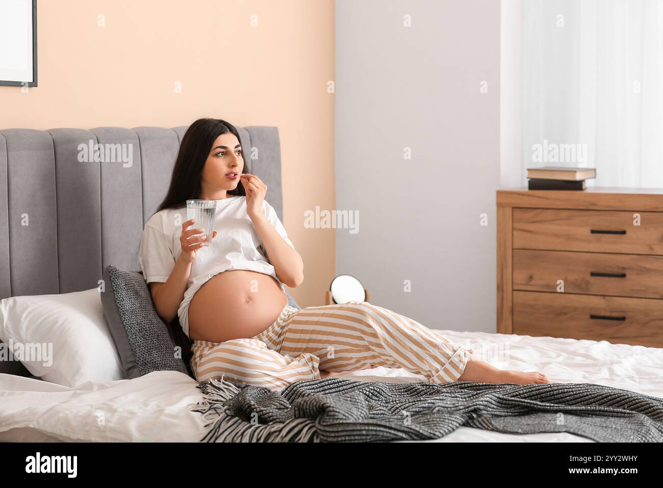 Bella donna incinta con un bicchiere d'acqua che prende pillole in camera da letto Foto Stock