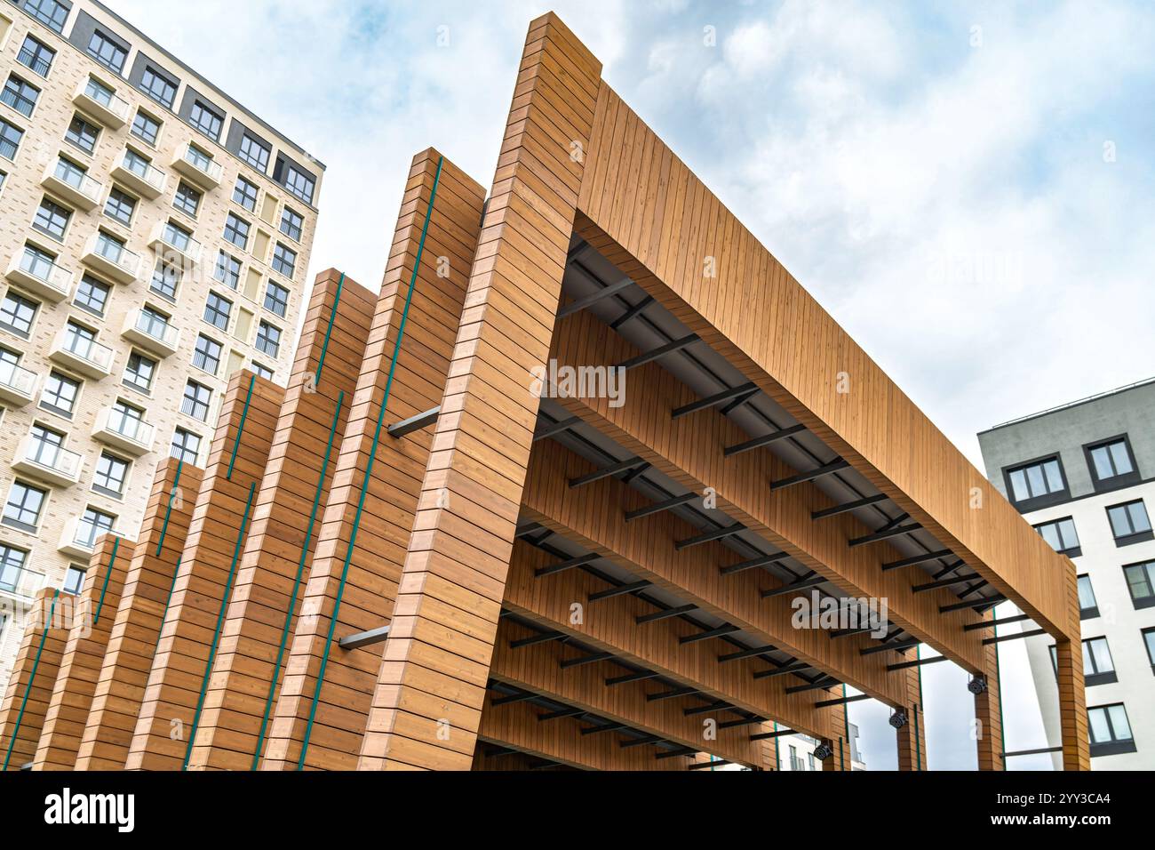 Moderno design architettonico in legno di palcoscenico all'aperto con travi sovrapposte e spazi aperti contro edifici residenziali e cielo blu nuvoloso. Concetto di Foto Stock