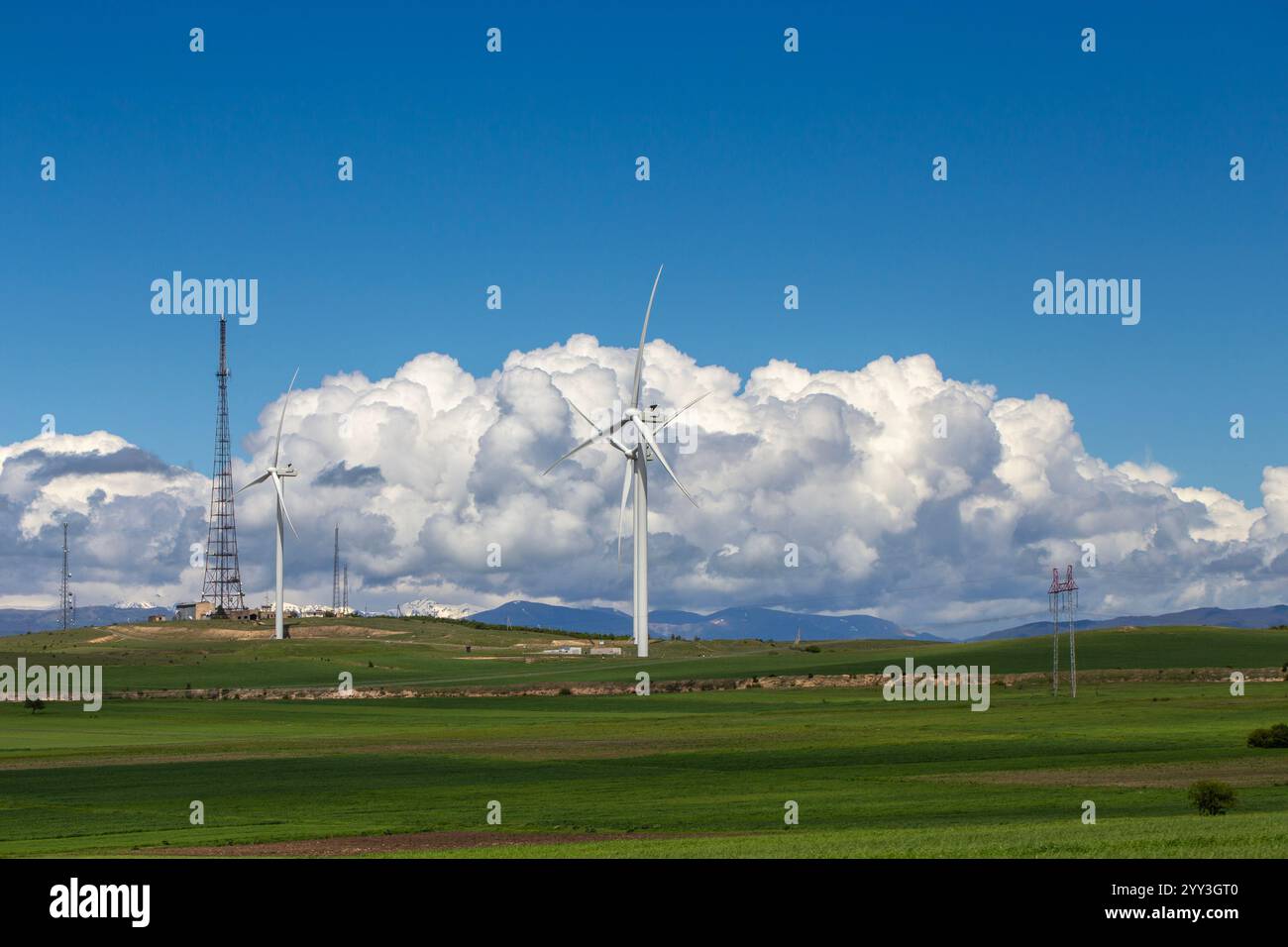 Un paesaggio georgiano panoramico caratterizzato da torreggianti turbine eoliche tra lussureggianti campi verdi sotto un cielo blu luminoso. Le montagne innevate si innalzano in lontananza Foto Stock