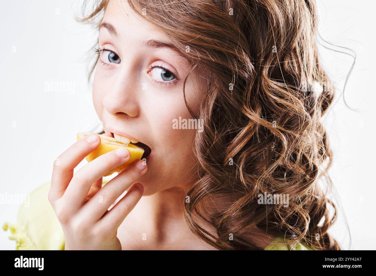 Una ragazza dai capelli ricci che muta una succosa fetta di limone, creando un viso esilarante in risposta all'intensa acidità, catturando l'essenza giocosa del bambino Foto Stock