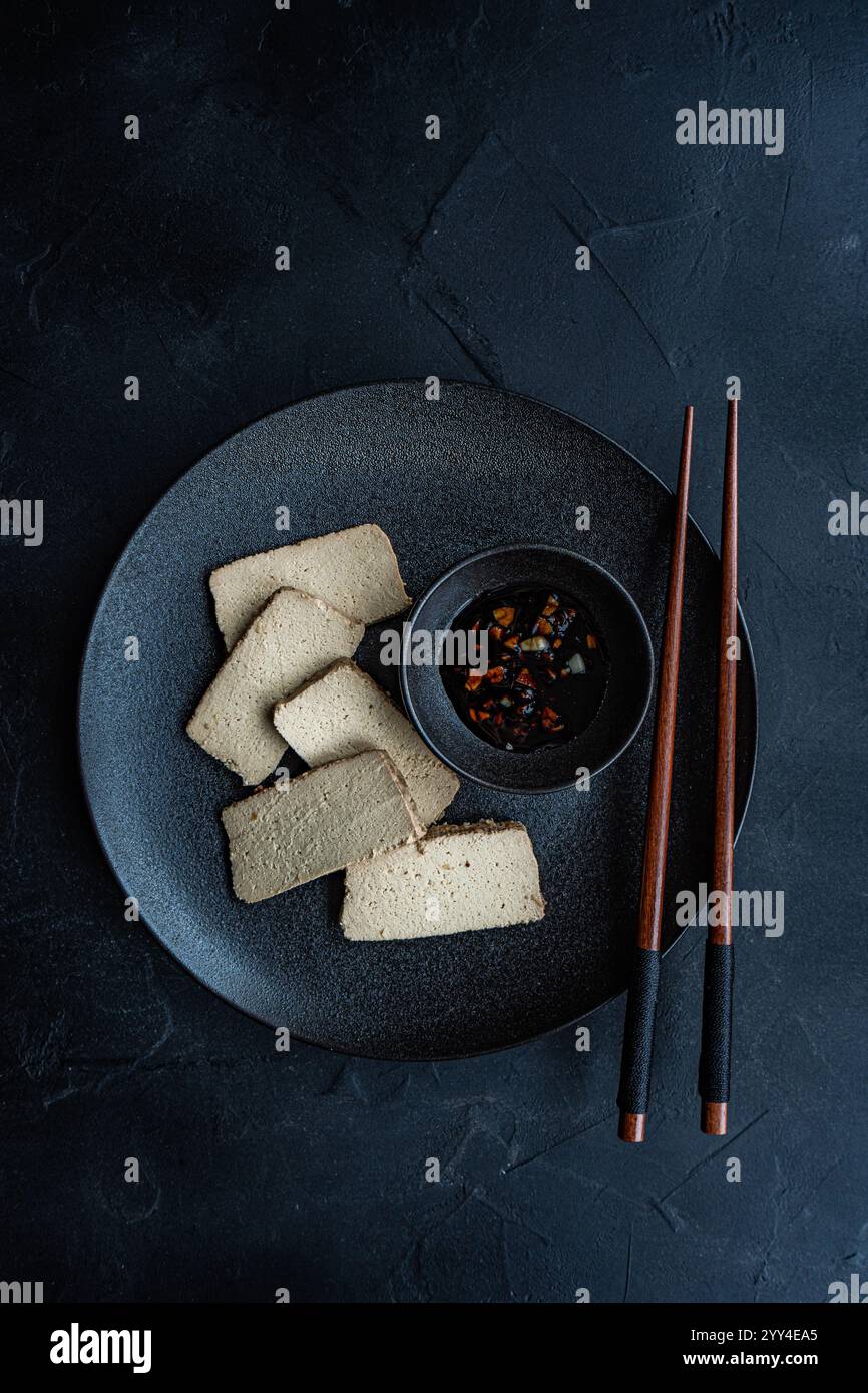Vista dall'alto delle fette di tofu al barbecue su un piatto scuro con salsa al pepe piccante e bacchette, servite su una superficie nera ruvida. Ideale per un sano pasto vegano Foto Stock