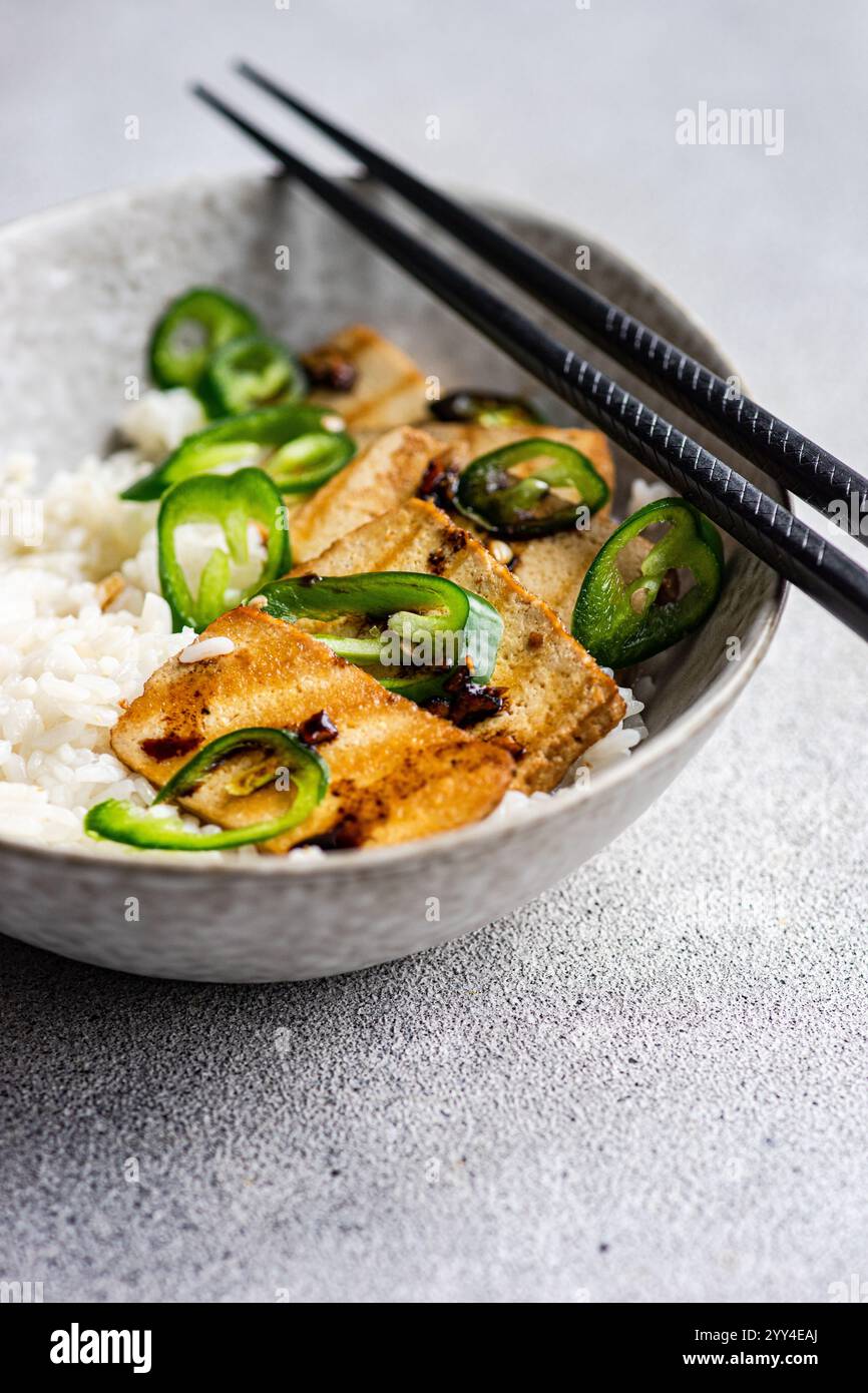 Da una vista angolata, questa immagine mostra una ciotola di fette di tofu barbecue servite su riso bianco, guarnite con fette di jalapeno verde fresco e accompagnato Foto Stock
