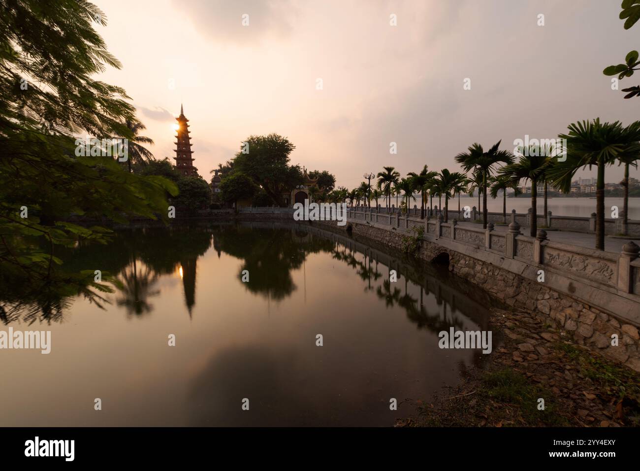 Un tranquillo tramonto su un tranquillo lago di Hanoi con un'iconica pagoda che si riflette nell'acqua le palme fiancheggiano il sentiero, offrendo una vista pittoresca di Foto Stock