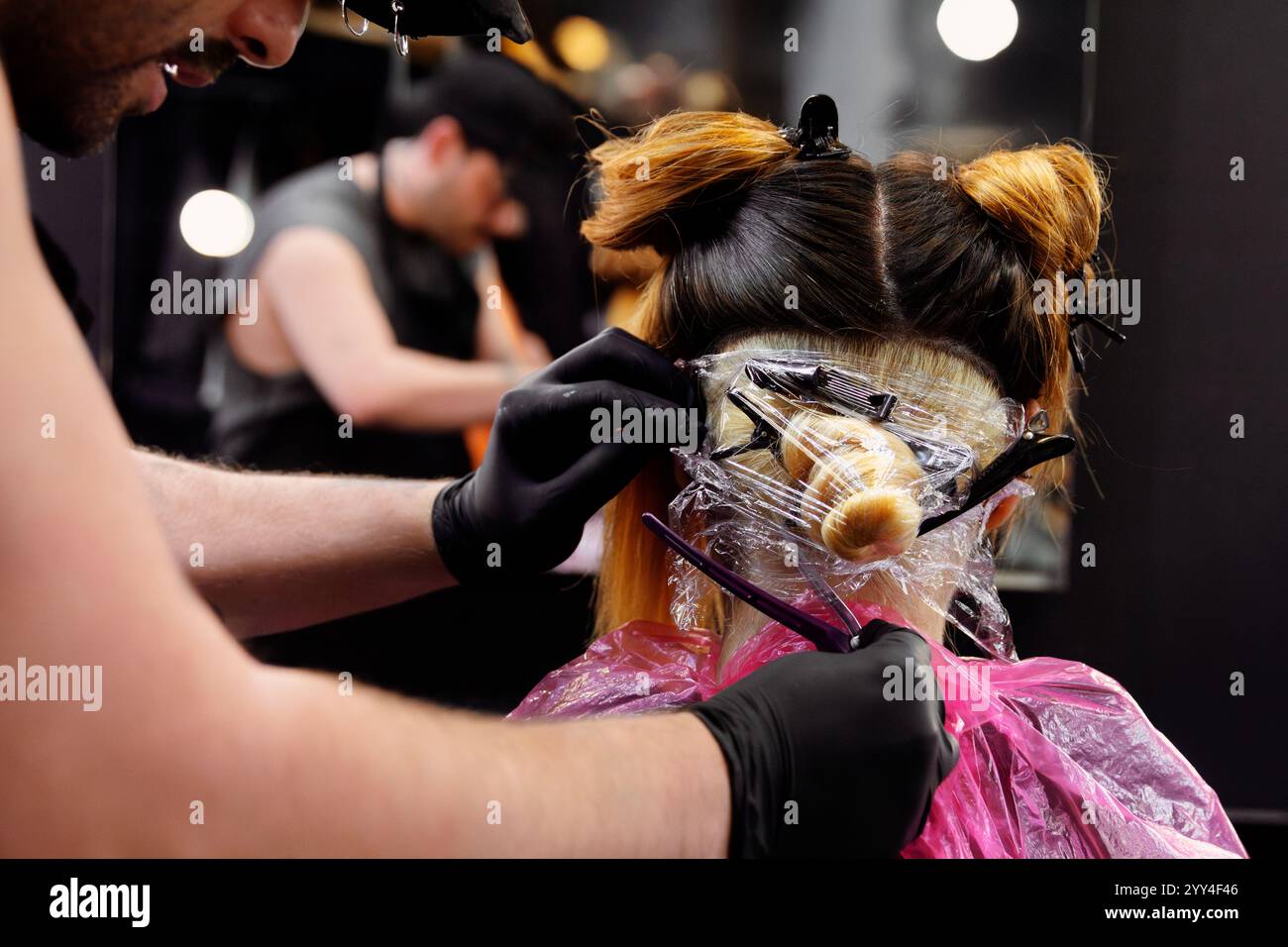 L'hair stylist che indossa i guanti applica il colore dei capelli ai capelli di un cliente avvolti in una pellicola di plastica in un salone la scena cattura un trattamento di bellezza professionale Foto Stock