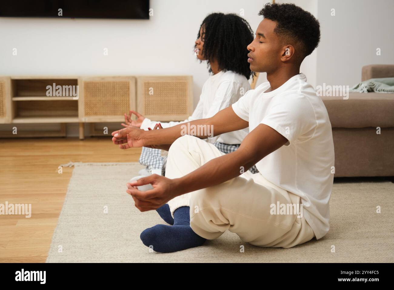 Una giovane coppia di neri gode di una tranquilla sessione di meditazione a casa, seduta a gambe incrociate sul pavimento in serena concentrazione, creando un atmos calmante Foto Stock