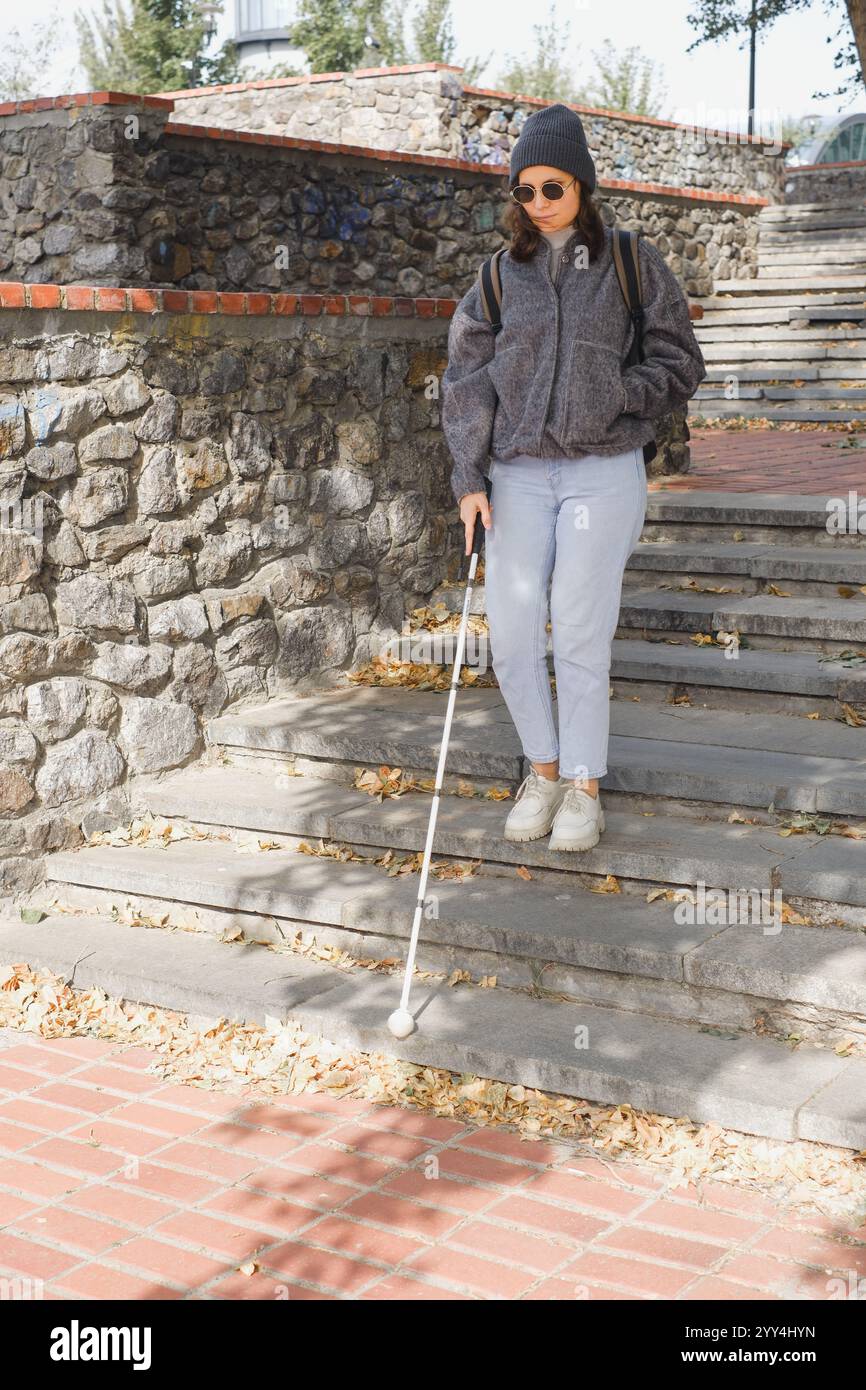 Donna cieca, con abbigliamento casual e occhiali da sole, naviga su una soleggiata scalinata all'aperto con il suo bastone bianco, simboleggiando l'indipendenza e l'autosufficienza Foto Stock