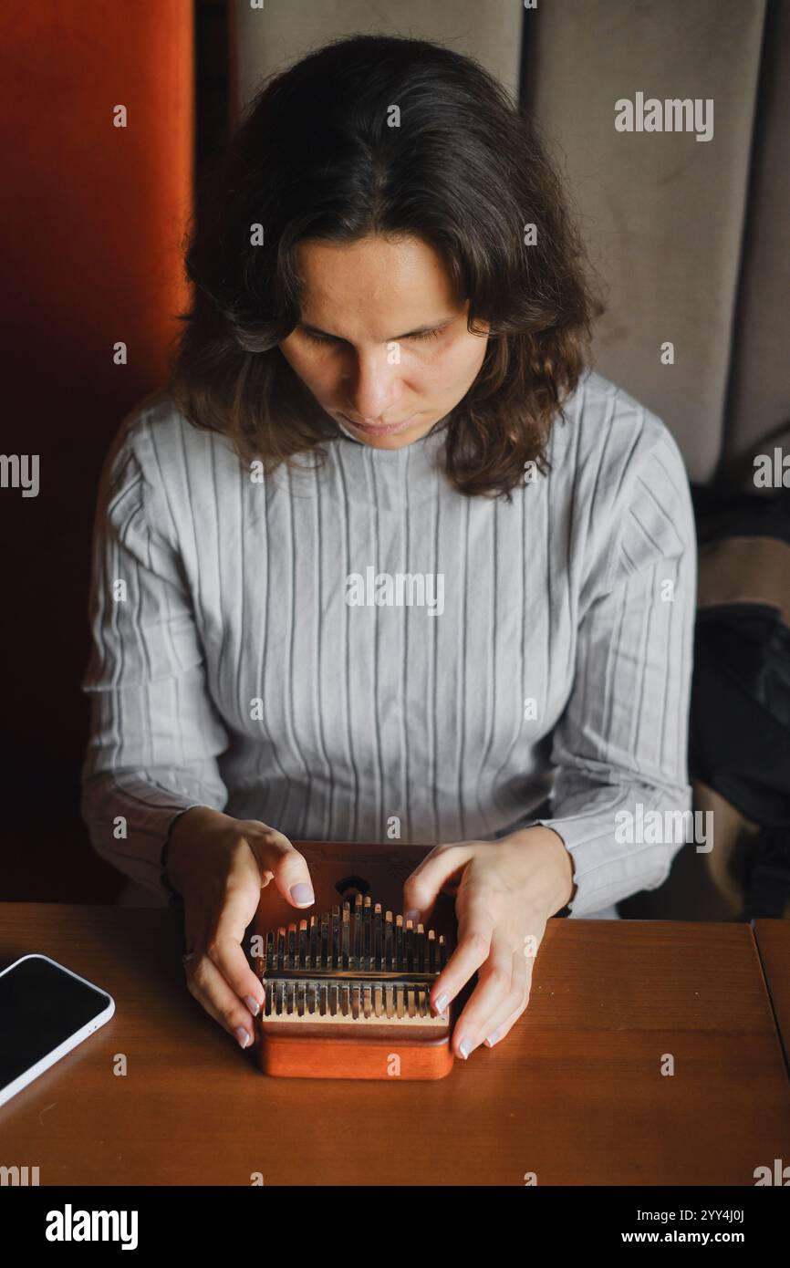 Una donna si concentra intensamente mentre le sue mani suonano abilmente un kalimba, un piccolo strumento a percussione in legno sul tavolo, uno smartphone, catturando una q Foto Stock