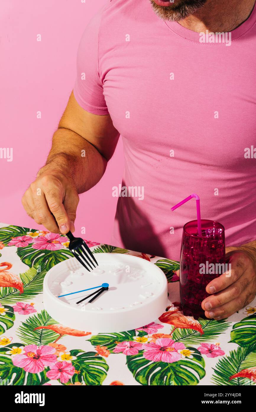 Una persona con una camicia rosa finge giocosamente di mangiare un orologio con una forchetta, simboleggiando l'ora dei pasti. Questa immagine è un'immagine a contrasto di una tovaglia floreale vivace Foto Stock