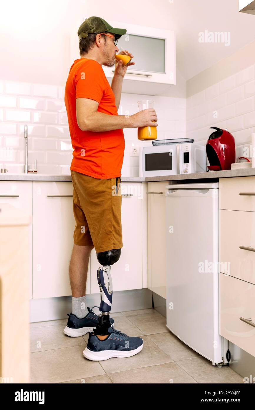 Un uomo con una gamba protesica si trova in una cucina moderna e beve succo d'arancia l'ambiente luminoso mostra la vita quotidiana con ausili ortopedici, enfasi Foto Stock