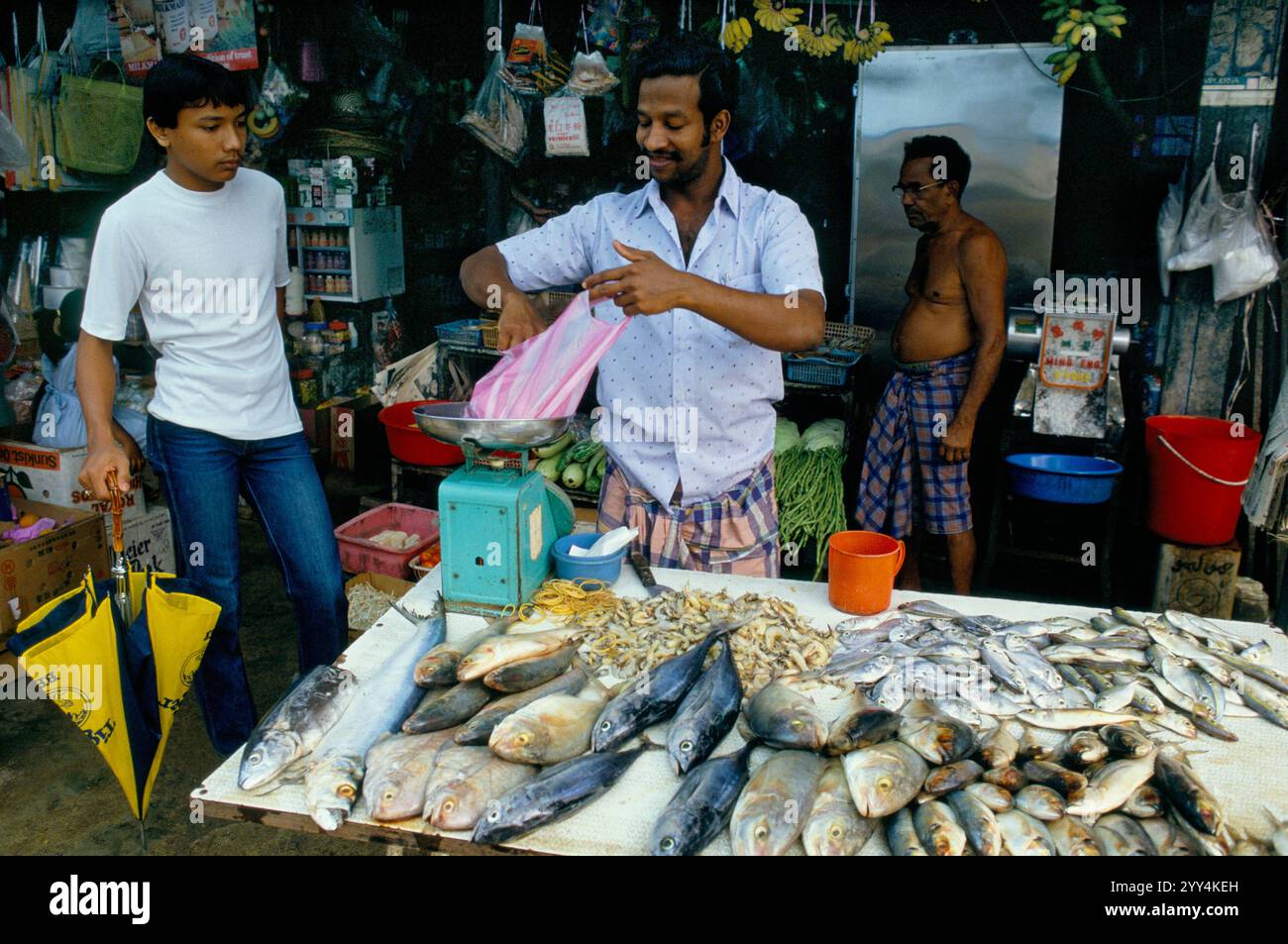 Singapore, negoziante che pesa un cliente che pesa il pesce che è appena stato acquistato. Kampong Fish Shop a Sembawang. Settembre 1983. Un kampong è un tipo di insediamento nei paesi del sud-est asiatico, queste abitazioni sono generalmente abbastanza basilari e potrebbero mancare di elettricità, acqua e gas. HOMER SYKES anni '1980 Foto Stock
