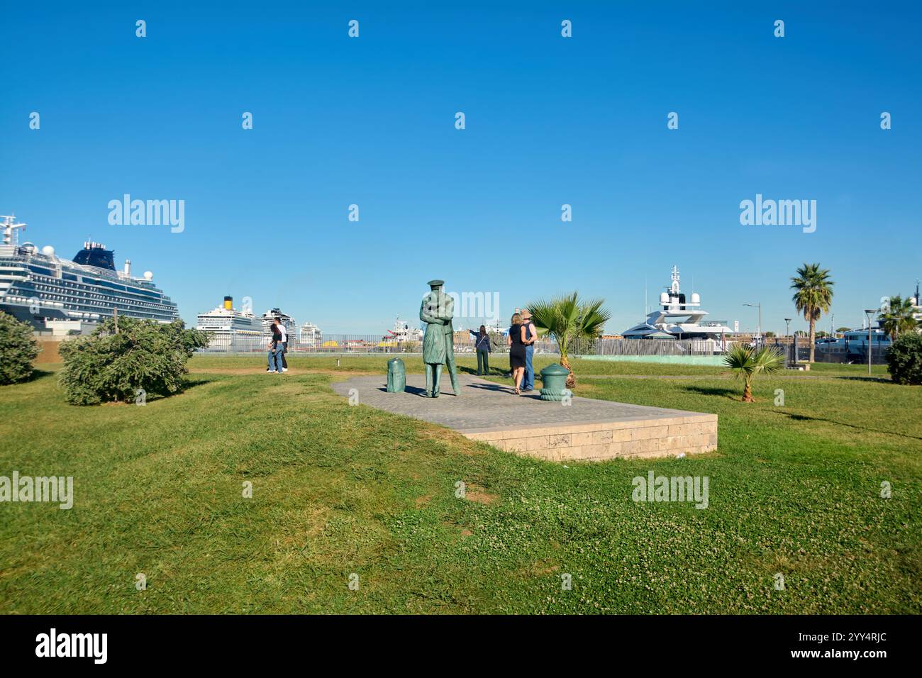 Civitavecchia, Italia - 19 dicembre 2024: Statua dell'ufficiale di marina nel parco sul lungomare e navi da crociera attraccate circondate da turisti che catturano lo scen Foto Stock