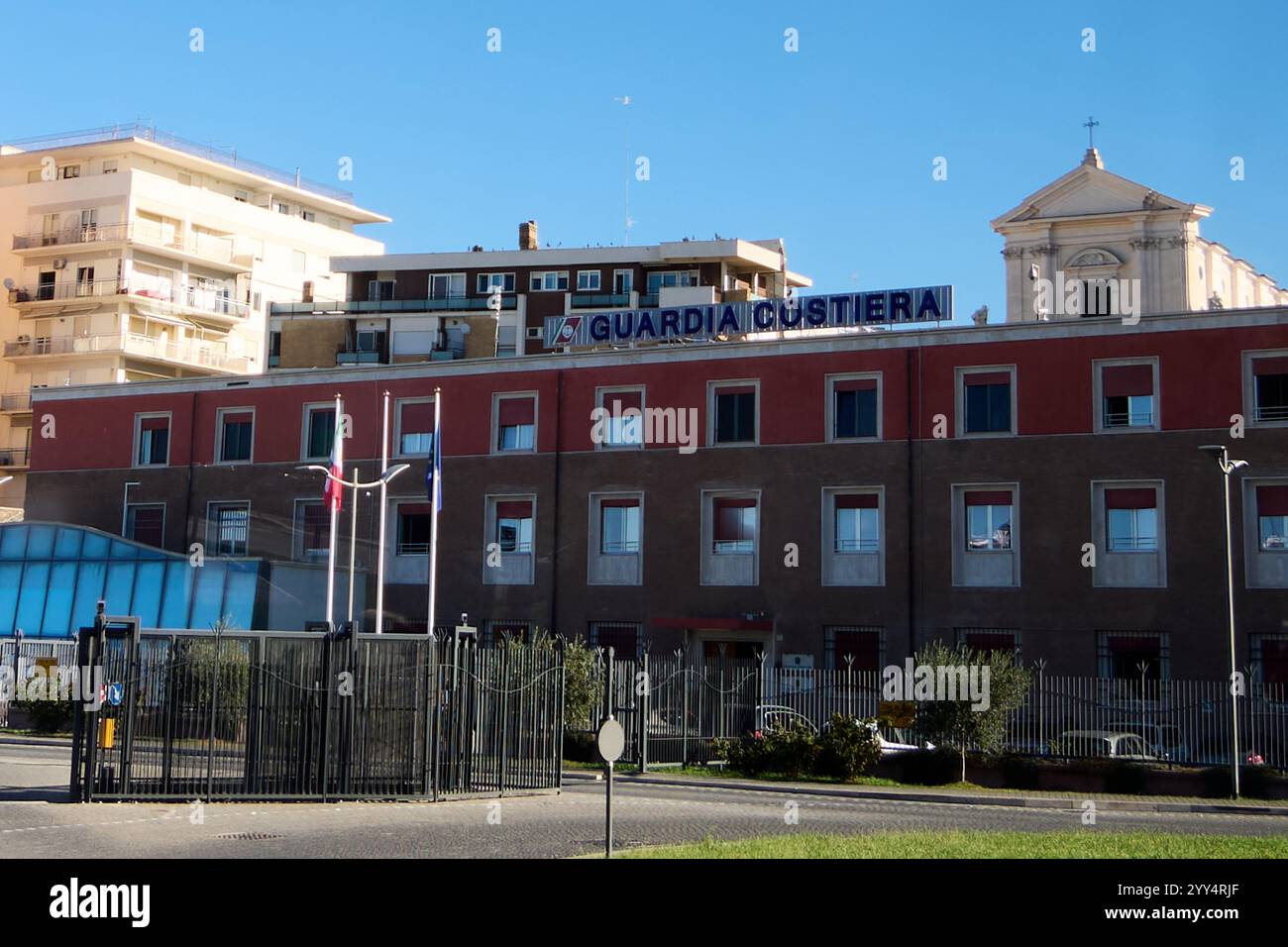 Civitavecchia, Italia - 19 dicembre 2024: Edificio della Guardia Costiera con finestre, bandiere e area recintata si erge di fronte ad una chiesa e ad un edificio residenziale Foto Stock