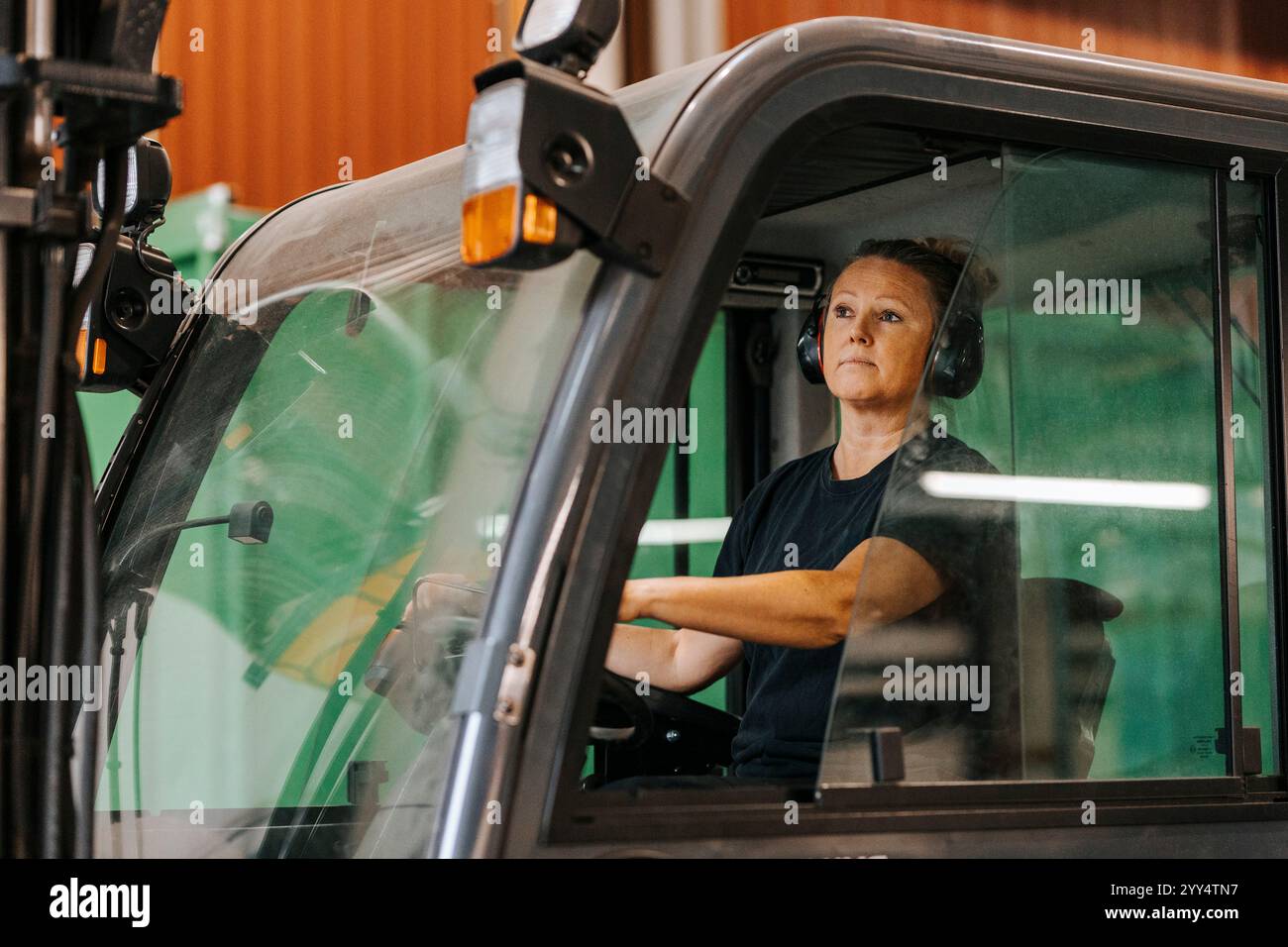 Lavoratrice matura che indossa le cuffie wireless mentre è seduta sul carrello elevatore in fabbrica Foto Stock