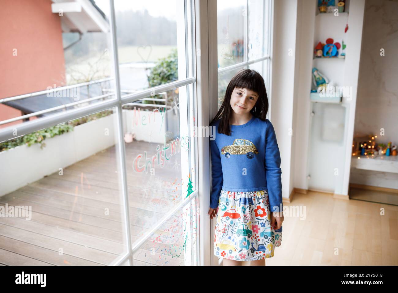 Ragazza a piedi nudi che indossa un abito colorato, che guarda i disegni natalizi sulla finestra, irradiando gioia e meraviglia durante le festività natalizie Foto Stock