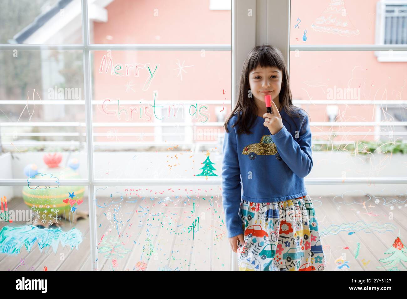 Ragazza a piedi nudi che indossa un abito colorato, che guarda i disegni natalizi sulla finestra, irradiando gioia e meraviglia durante le festività natalizie Foto Stock