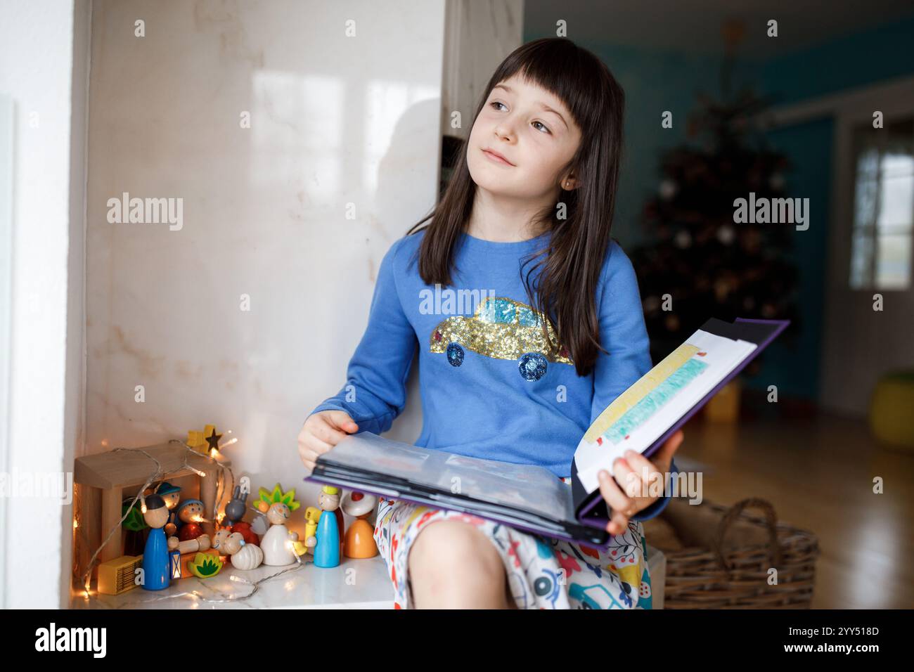 Ragazza a piedi nudi che indossa un abito colorato, che guarda i disegni natalizi sulla finestra, irradiando gioia e meraviglia durante le festività natalizie Foto Stock