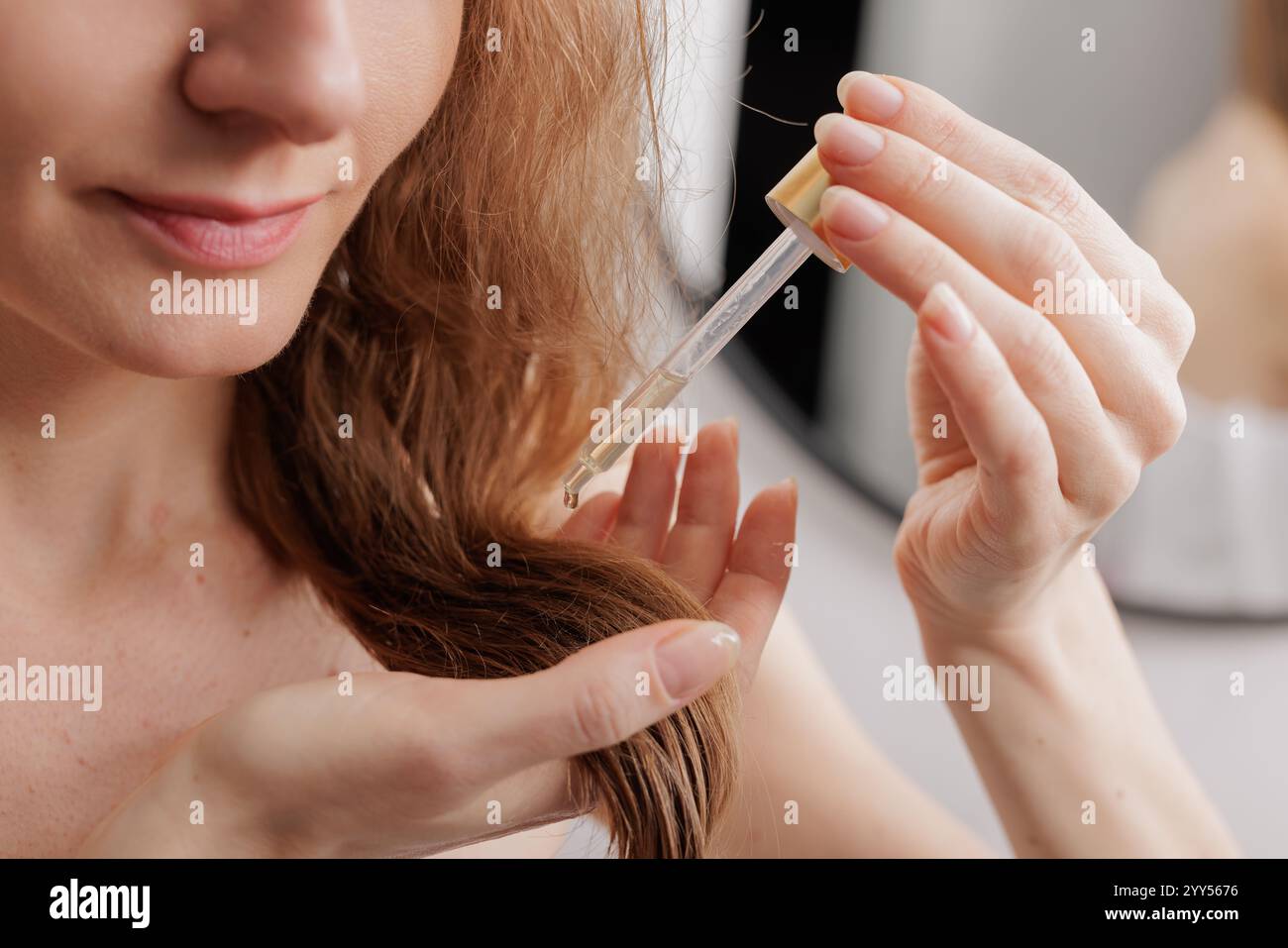 Tagliata di donna bionda che applica olio sui capelli mentre si acconciano i suoi bellissimi capelli in bagno dopo la doccia del mattino, gustando un nuovo prodotto per la cura dei capelli, c Foto Stock
