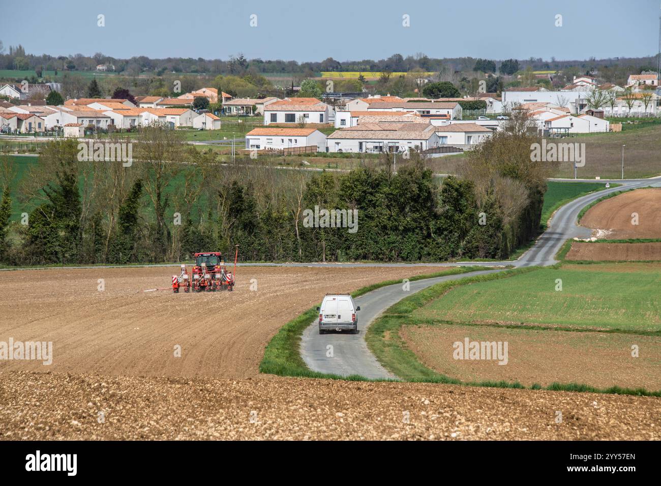Saint-Savinien (Francia centro-occidentale): Auto che passa su una strada di campagna vicino a un complesso abitativo e trattore con seminatrice in un campo Foto Stock