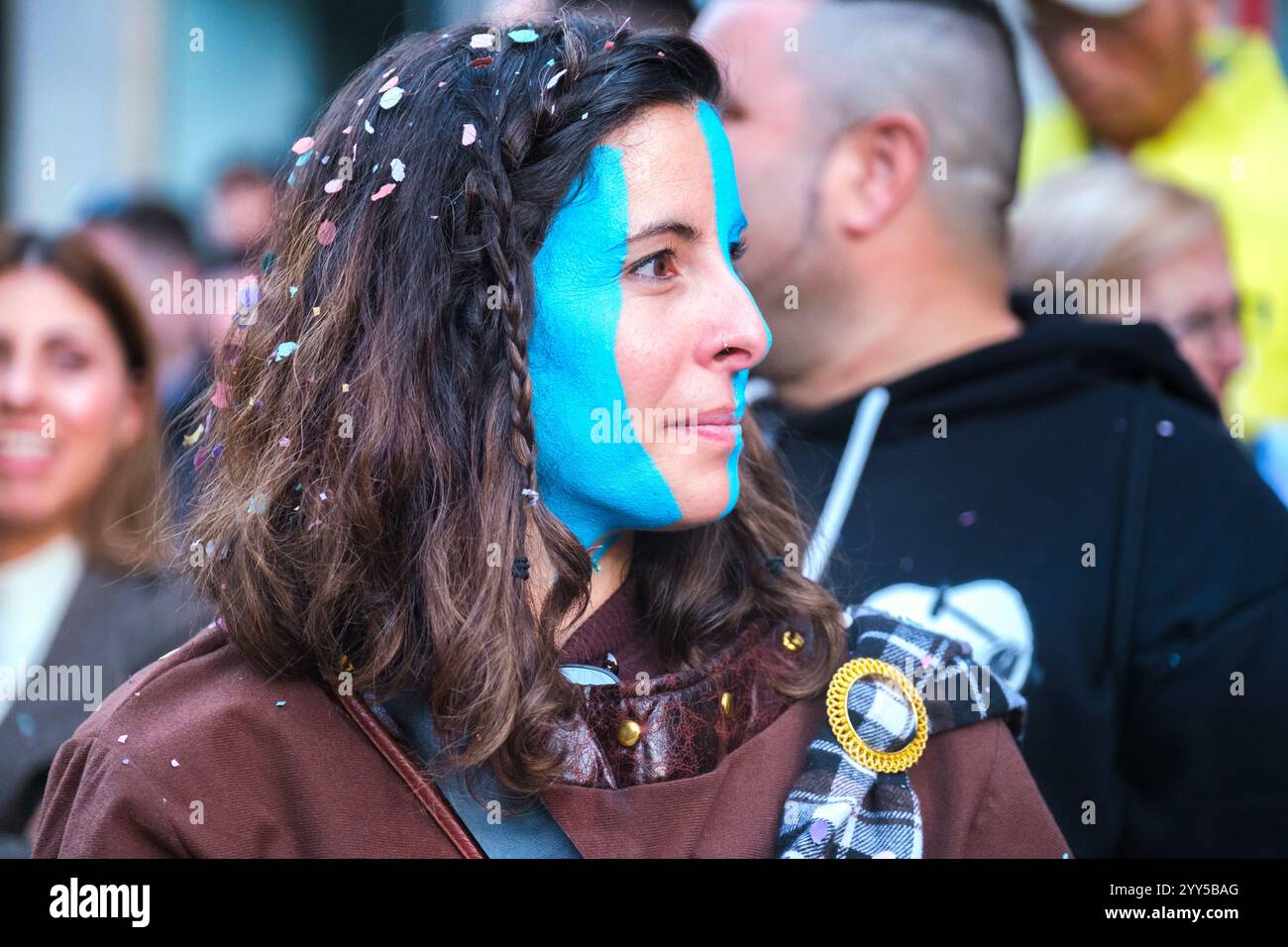 Persona all'evento all'aperto con mezza faccia dipinta in modo creativo di blu brillante, suggerendo una celebrazione o una causa, macchiata in una giacca marrone vivace. Foto Stock