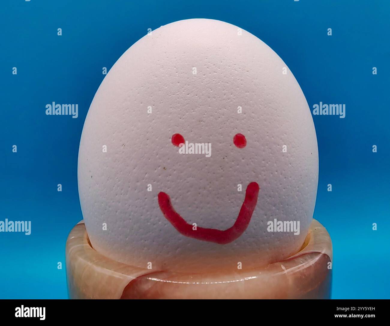Gioco creativo. Uovo bianco con sorriso Happy Face in Mini tazza marrone di marmo - Un'esposizione divertente e stravagante. Isolato su sfondo di carta blu Foto Stock