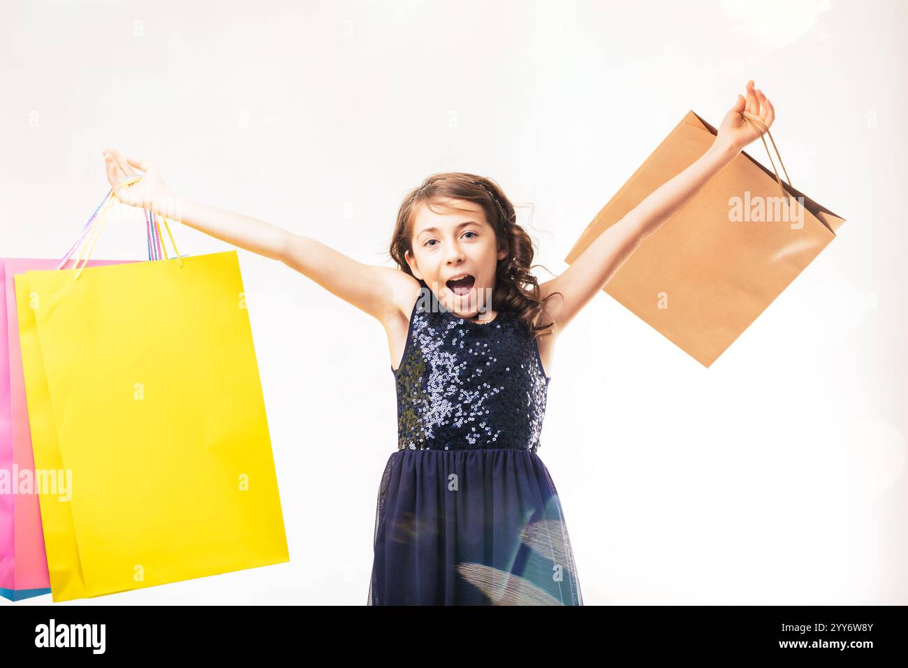 Eccitata ragazza elementare che urla con gioia mentre tiene in mano borse colorate, braccia allungate per festeggiare, isolata contro un bianco pulito Foto Stock