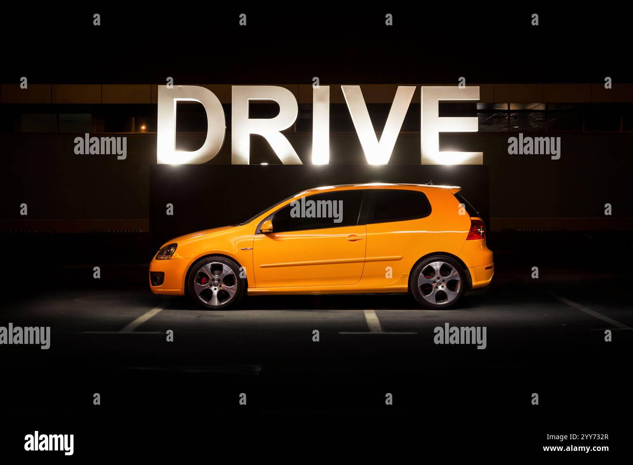 Orange Volkswagen Golf di notte vicino al cartello 'DRIVE'. Vista laterale del berlina Volkswagen GTi in un parcheggio buio. Foto Stock