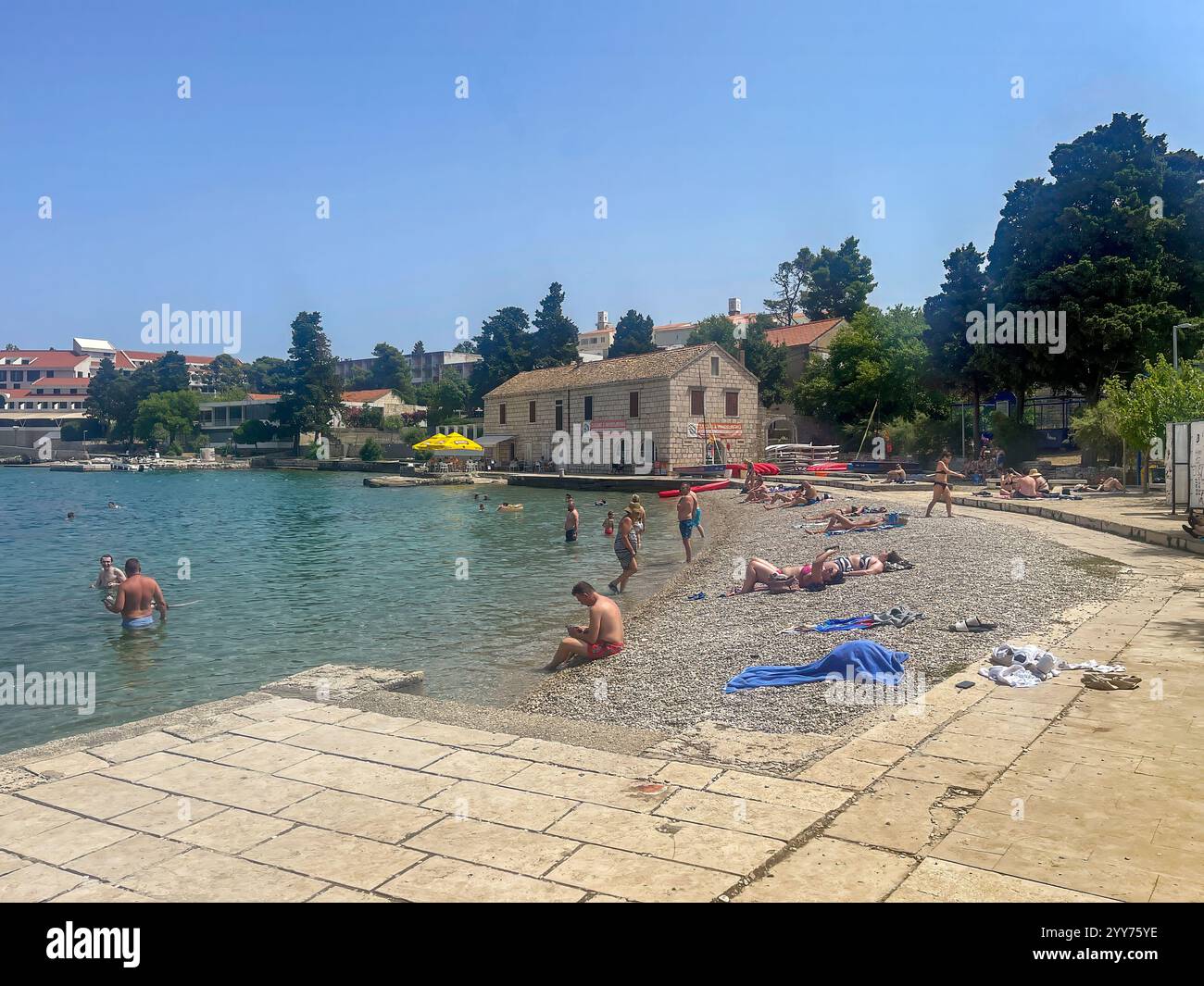 Korcula, Croazia - 30 giugno 2024: Spiaggia di Banje e Oreb Club International Sailing & Windsurf School Building. Bagnanti in mare e sui ciottoli Foto Stock