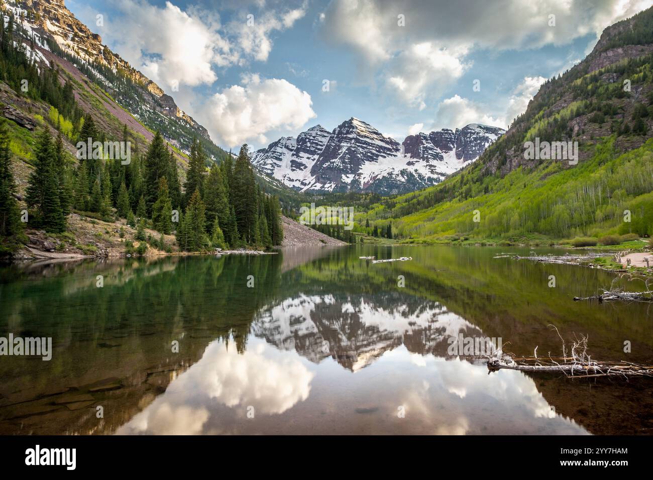 Le splendide montagne Maroon Bells si innalzano sopra un tranquillo lago, circondate da vegetazione lussureggiante e vette innevate sotto un cielo blu brillante. Foto Stock