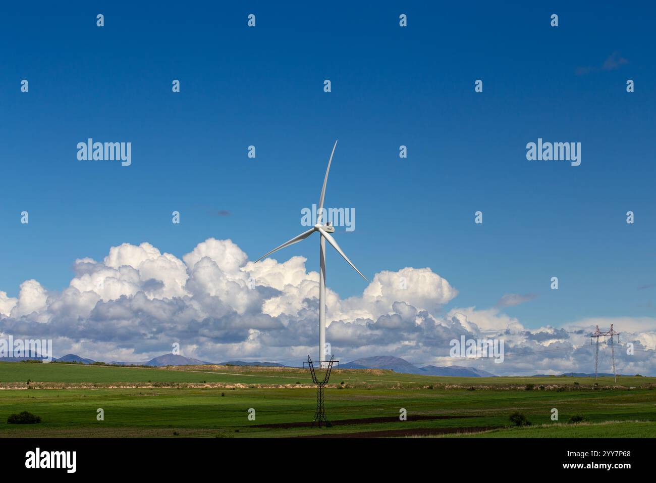 Un paesaggio georgiano panoramico caratterizzato da torreggianti turbine eoliche tra lussureggianti campi verdi sotto un cielo blu luminoso. Le montagne innevate si innalzano in lontananza Foto Stock