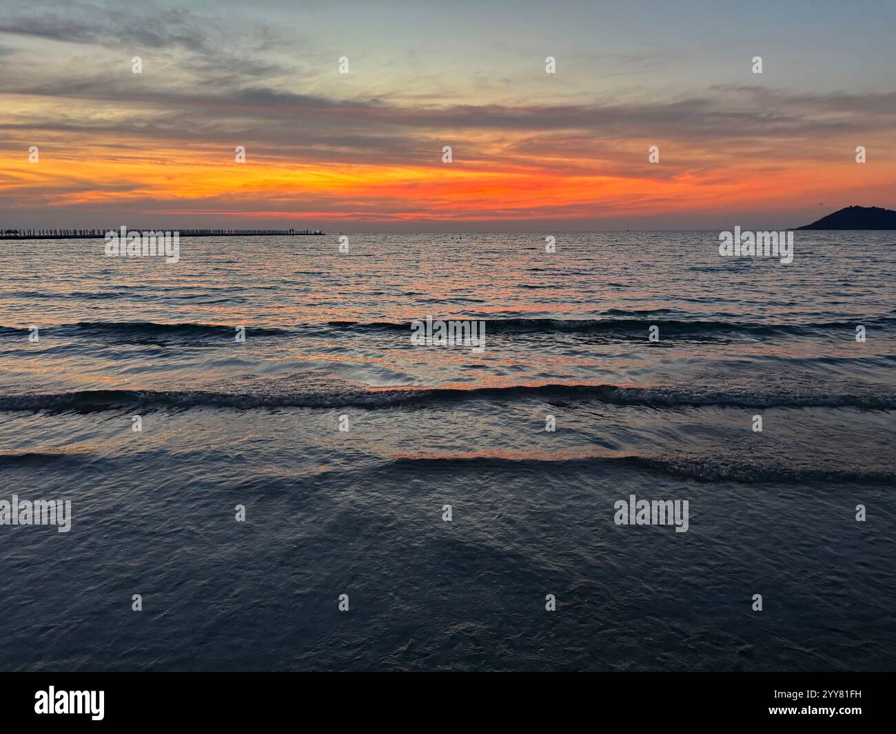 Splendido tramonto arancione su una spiaggia con onde calme e un lungo molo - Immagine stock catturata con smartphone