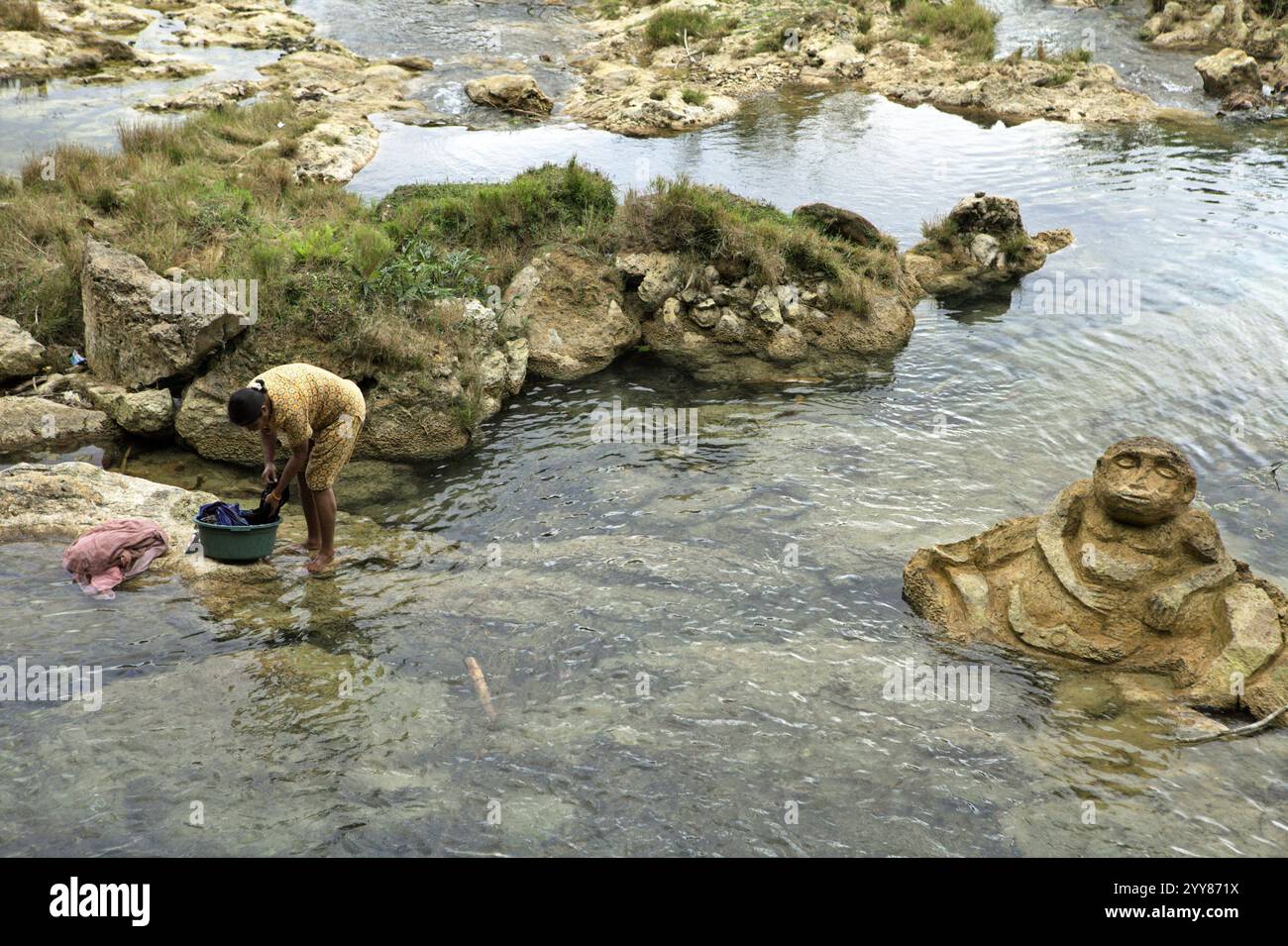 Una donna che lava vestiti su un ruscello vicino a una pietra scolpita di figura umana vicino a Waikelo Sawah, una rara fonte d'acqua nell'isola di Sumba, Indonesia. Foto Stock