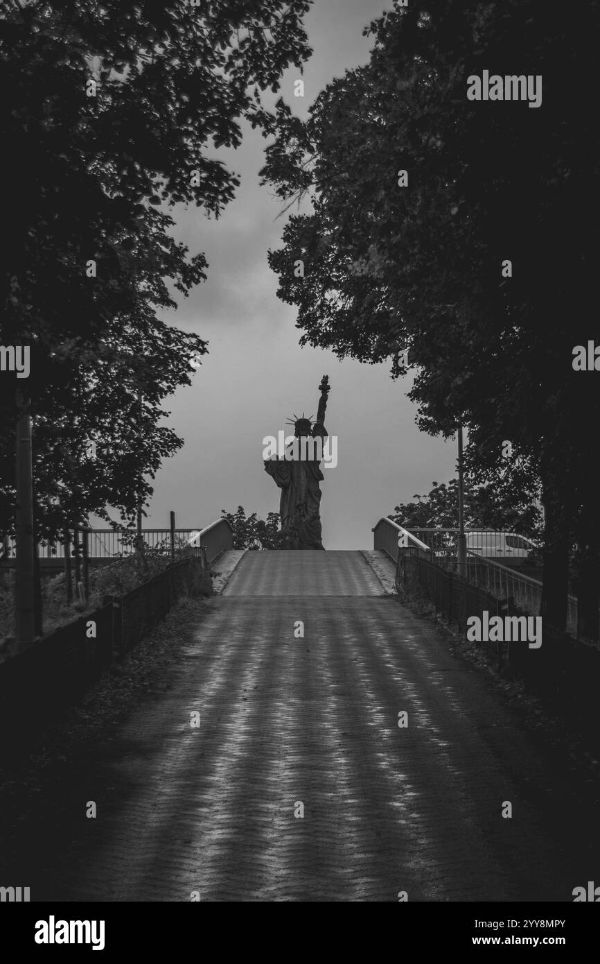 Un percorso piovoso conduce a una replica della Statua della libertà, incorniciata da alberi e nuvole, catturando un momento sereno ma drammatico Foto Stock
