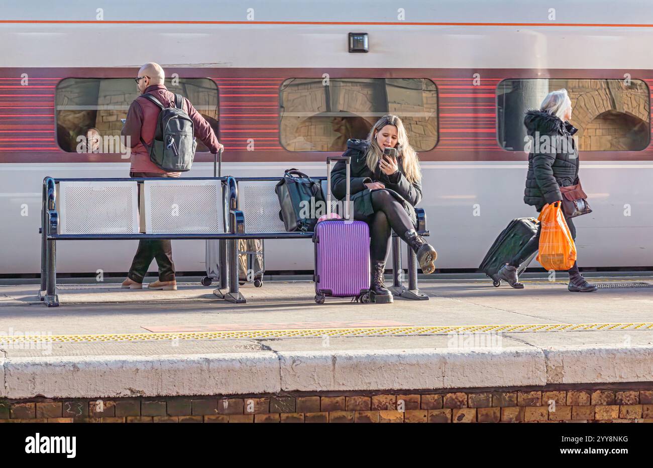Tre passeggeri sono su una piattaforma della stazione ferroviaria. Uno è seduto su una panchina, uno sta in piedi e un altro cammina. Una carrozza ferroviaria è sullo sfondo. Foto Stock