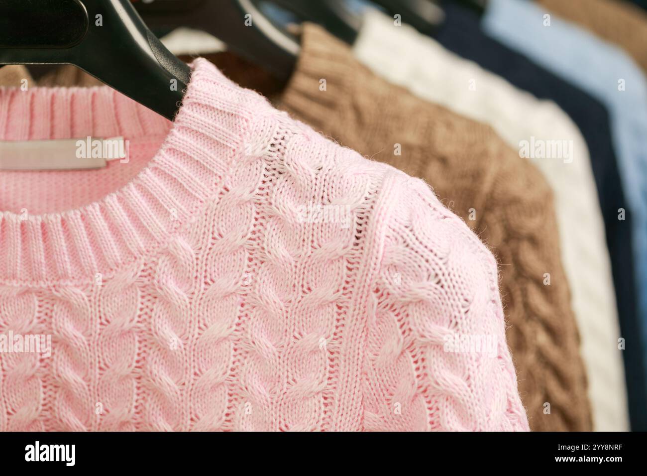 Uno scatto ravvicinato di un maglione in maglia a cavo rosa pastello esposto su un appendiabiti, allestito tra gli altri capi dalle tonalità neutre in un negozio di abbigliamento, evidenziandolo Foto Stock