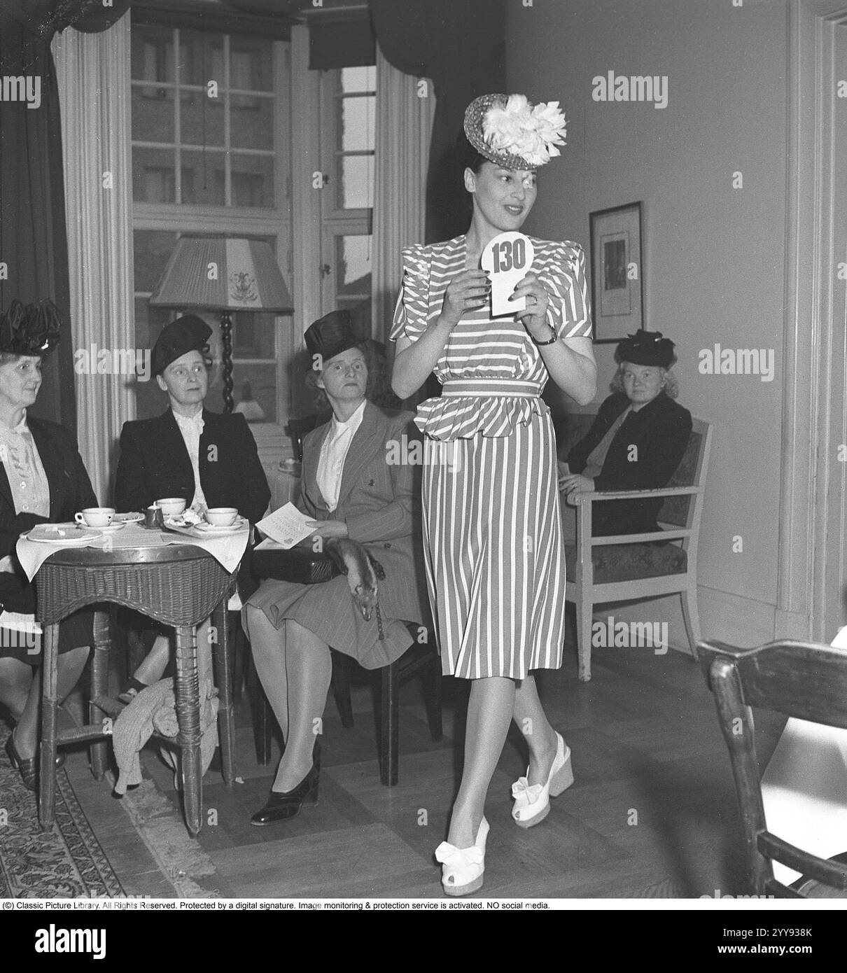 Sfilata di moda 1946. Una giovane modella dai capelli scuri ad una sfilata di moda. Indossa un abito anni '1940 abbinato a una gonna a righe e a una giacca in vita. Cammina con sicurezza con scarpe bianche con tacco alto e un cappello intelligente, mostrando i vestiti alle donne che guardano. 1946 Kristoffersson rif. T148-4 Foto Stock