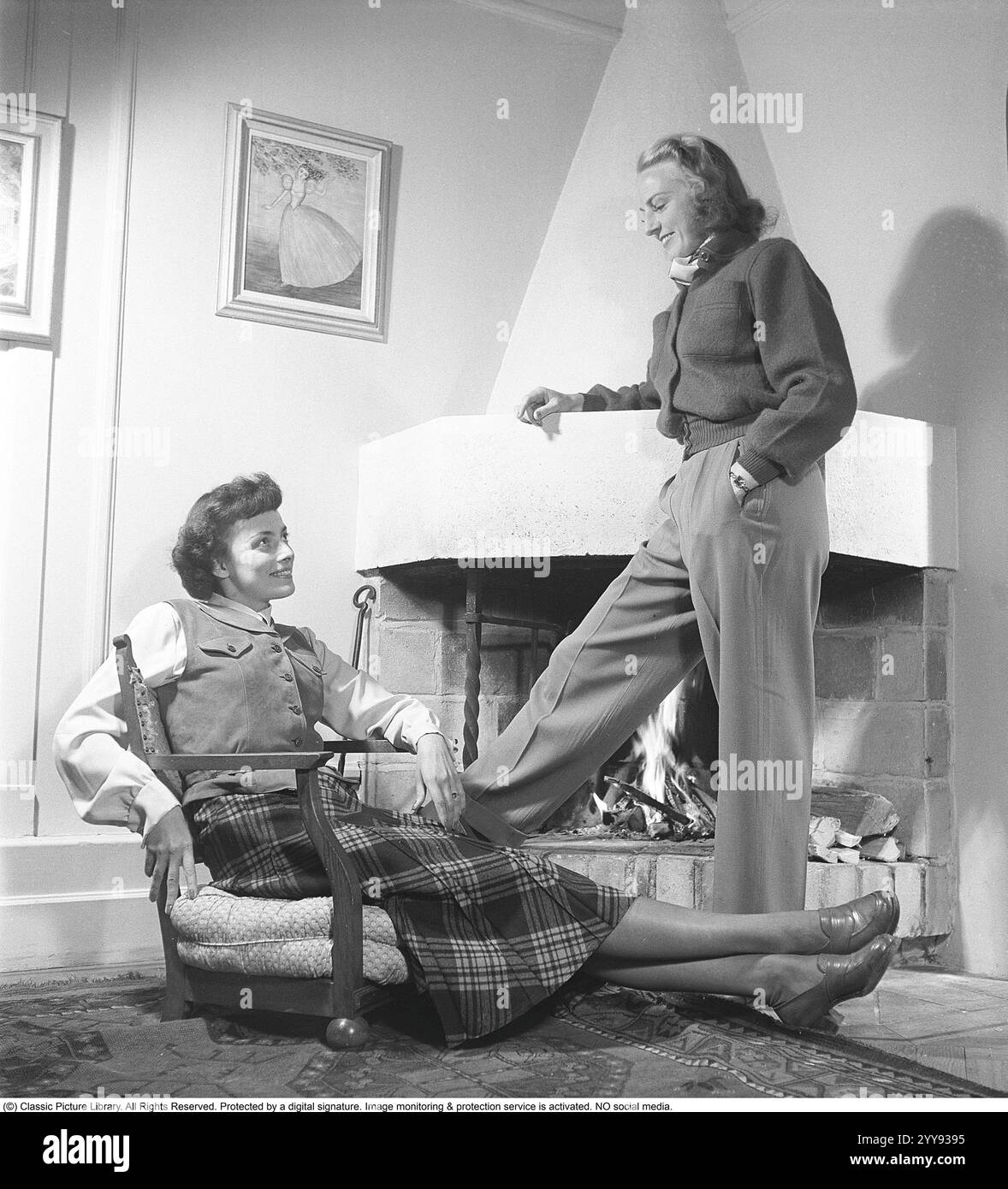 Alla moda negli anni '1940 Due donne in una stanza dove con un fuoco che brucia nel caminetto. Uno seduto in una poltrona dall'aspetto straordinario che non ha gambe. Entrambi indossano i tipici vestiti e la moda degli anni '1940. . Kristoffersson rif. AR116-1 Foto Stock