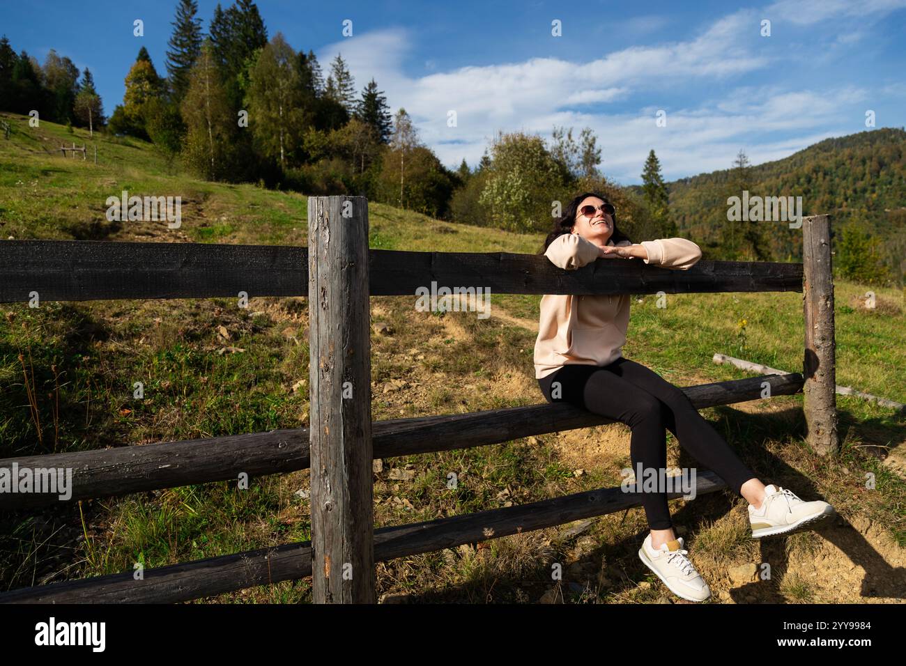 Giovane donna dai capelli ricci che indossa occhiali da sole seduti su una recinzione di legno in uno splendido paesaggio montano, abbracciando la libertà nelle lussureggianti foreste e soleggiato sk Foto Stock