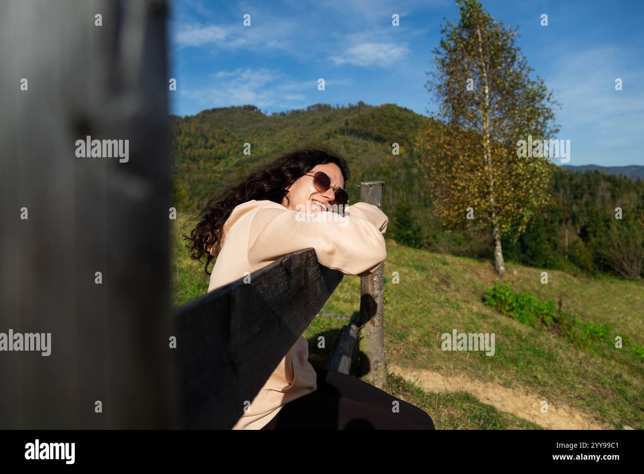 Giovane donna dai capelli ricci che indossa occhiali da sole seduti su una recinzione di legno in uno splendido paesaggio montano, abbracciando la libertà nelle lussureggianti foreste e soleggiato sk Foto Stock