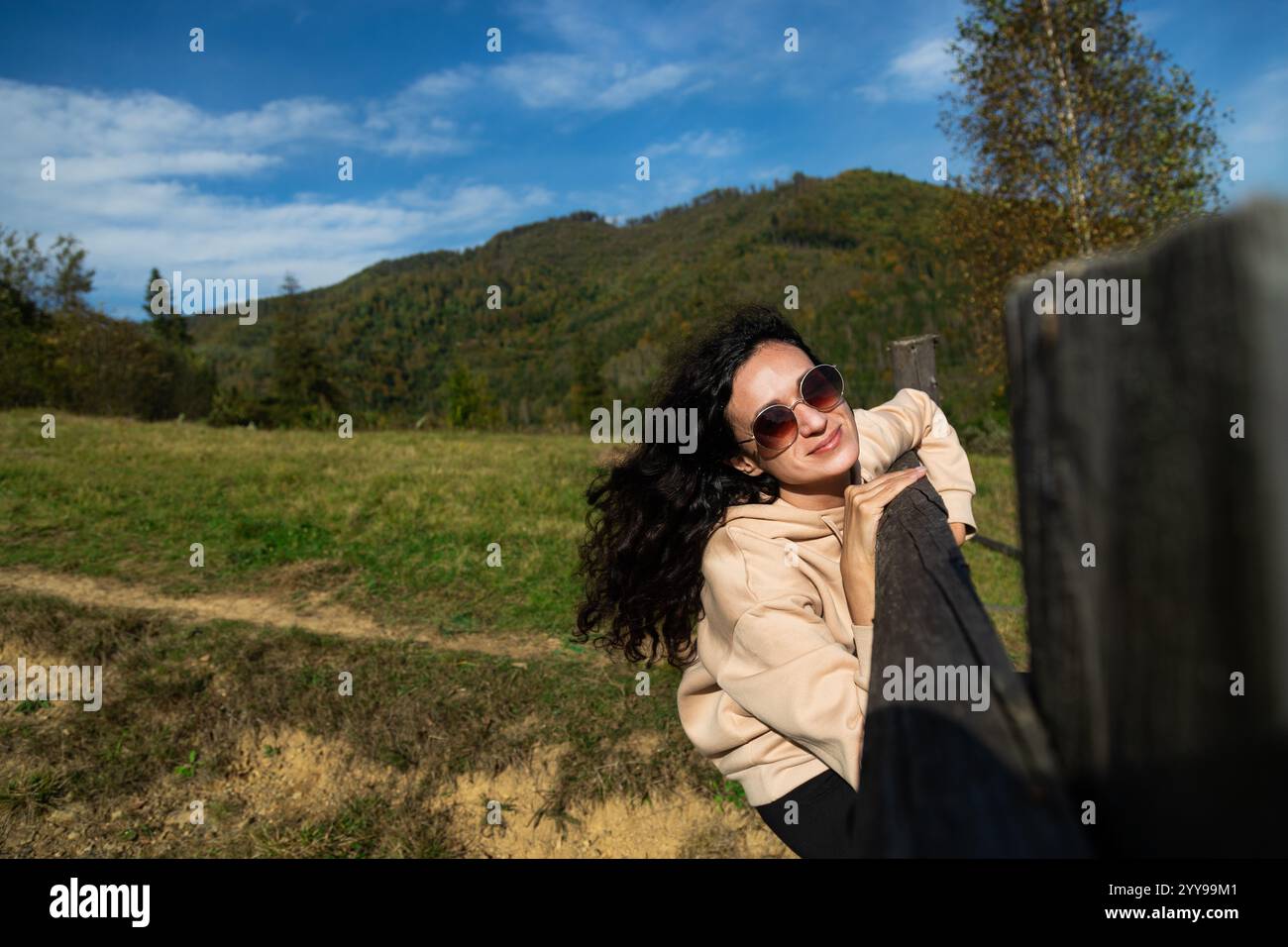 Giovane donna dai capelli ricci che indossa occhiali da sole seduti su una recinzione di legno in uno splendido paesaggio montano, abbracciando la libertà nelle lussureggianti foreste e soleggiato sk Foto Stock